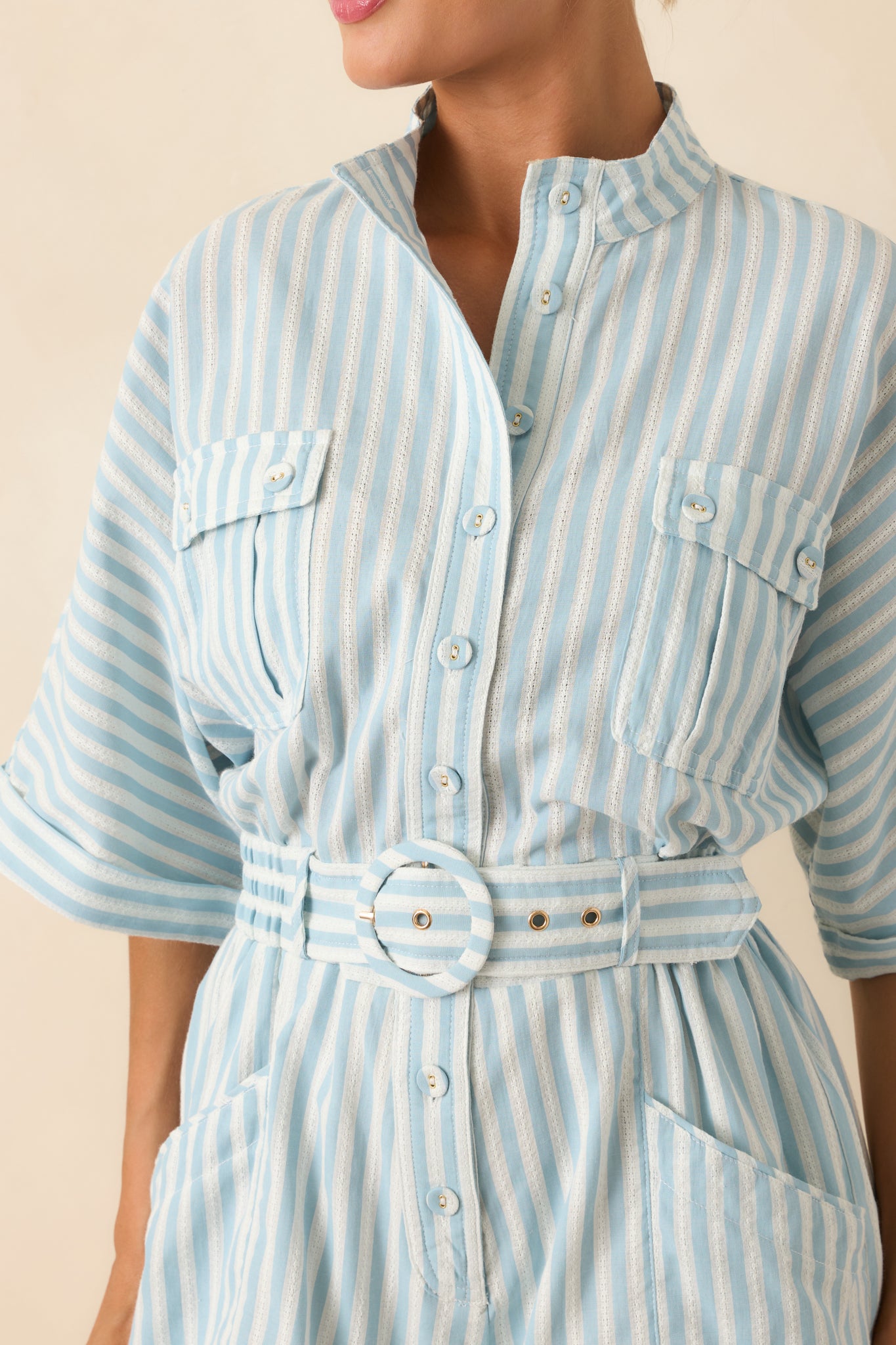 Detail close-up photo of light blue striped romper featuring detailed stitching, collared neckline, button front, cuffed short sleeves, and belted waist.