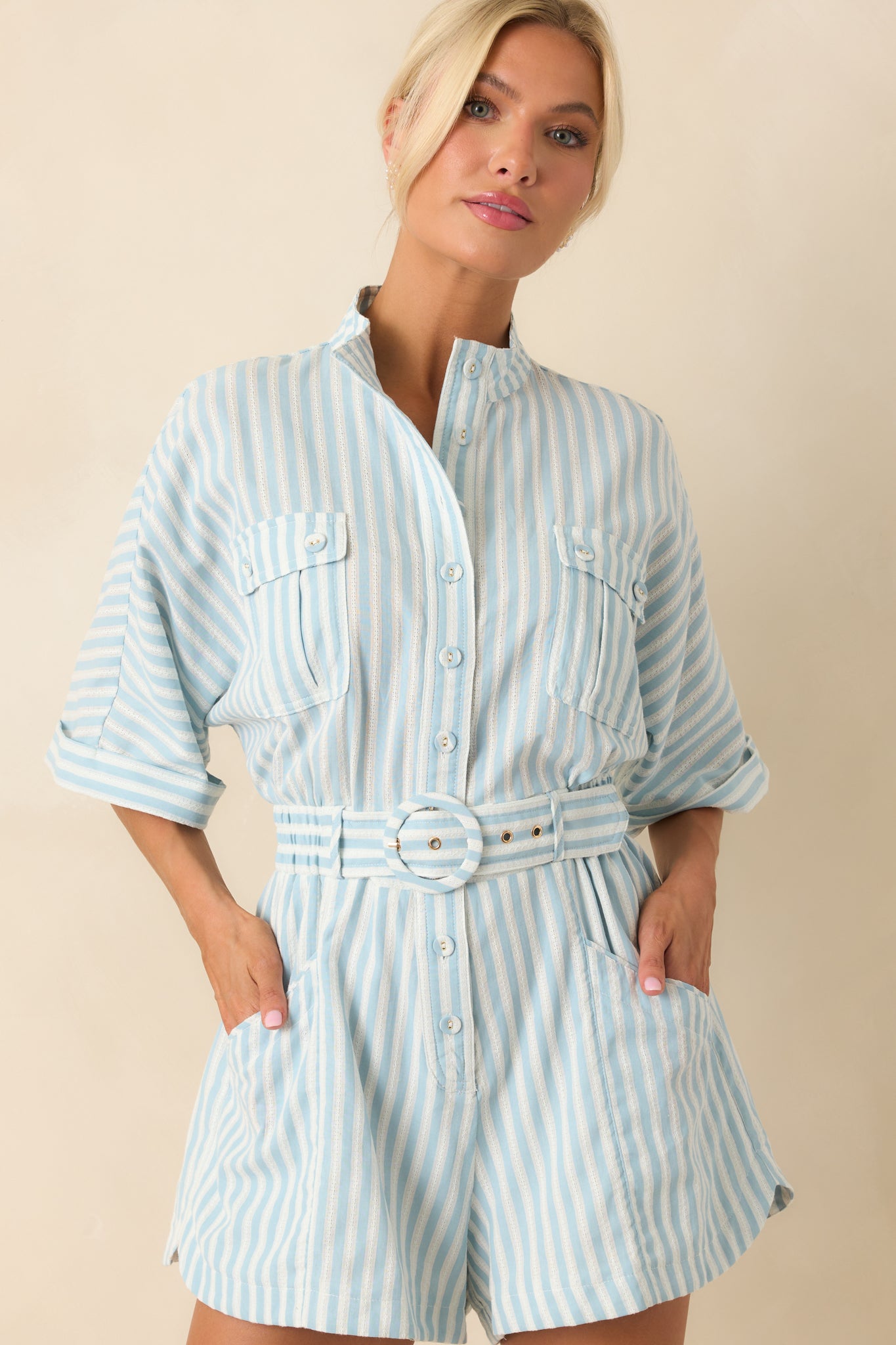Close-up of Prosperina light blue striped romper featuring detailed stitching, collared neckline, button front, cuffed short sleeves, belted waist, and front patch pockets.