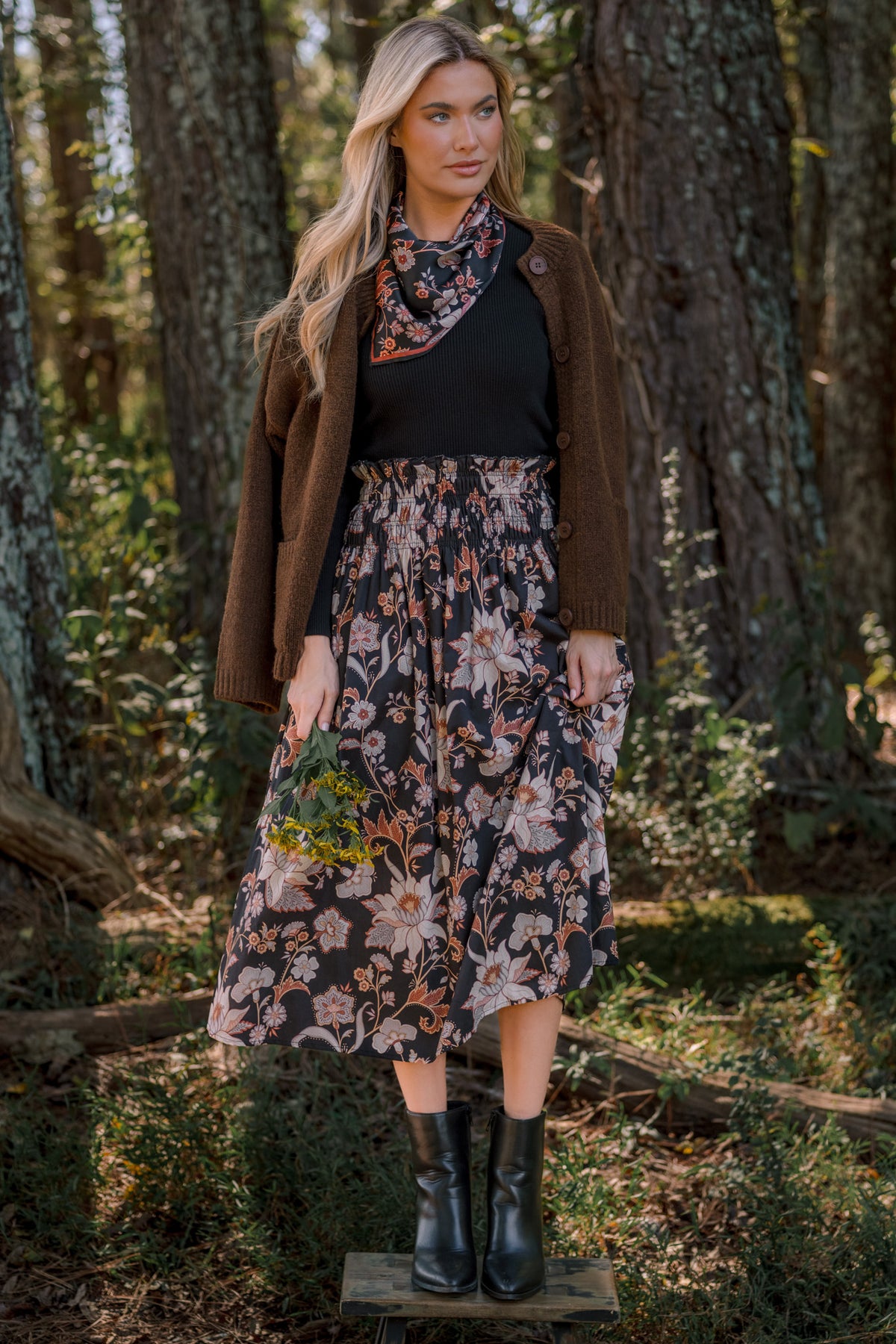 Model wearing a brown button-front cardigan over a black top with a floral midi skirt and black boots outdoors