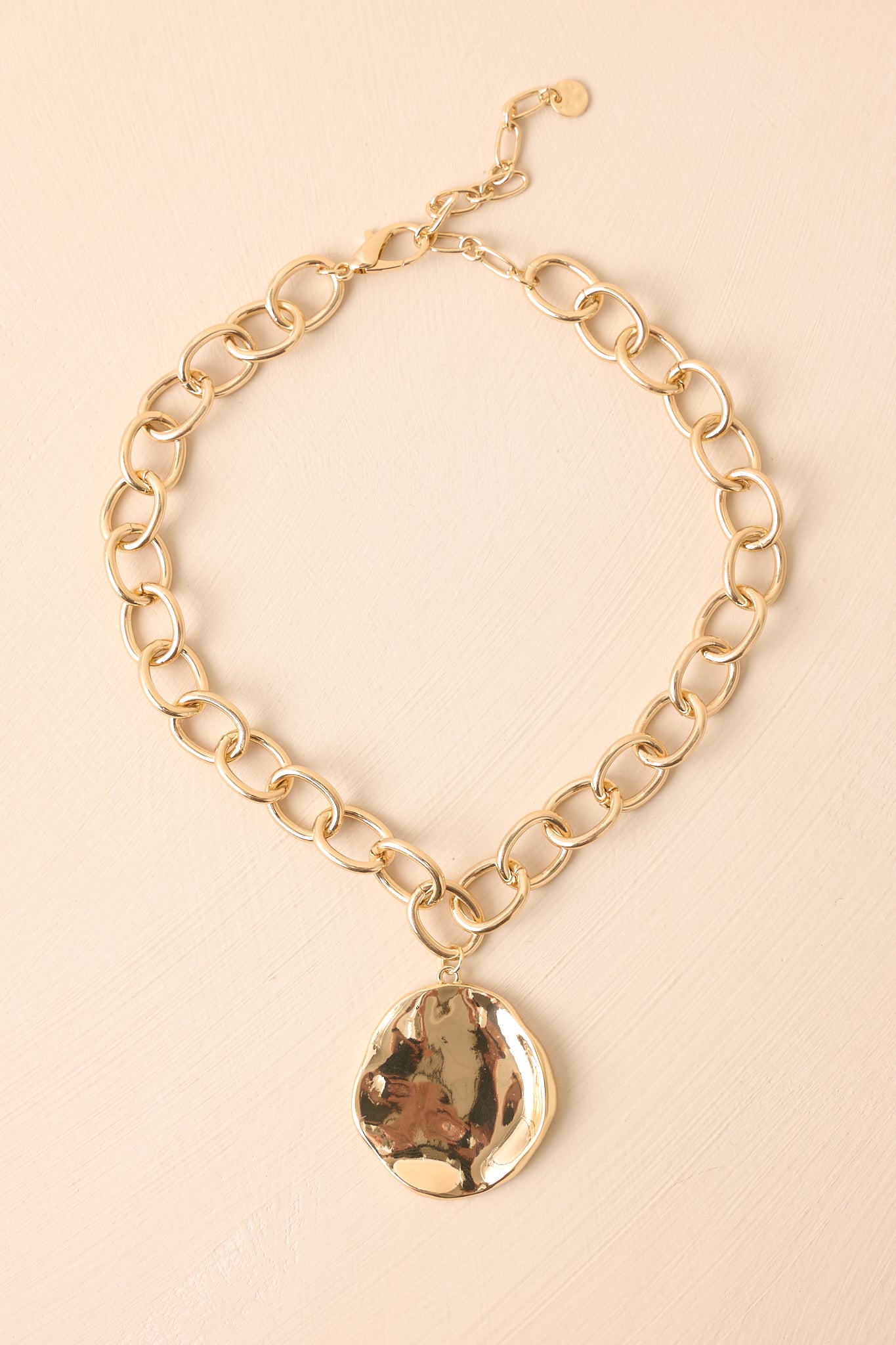 Gold necklace with abstract hammered pendant resting against a beige background.