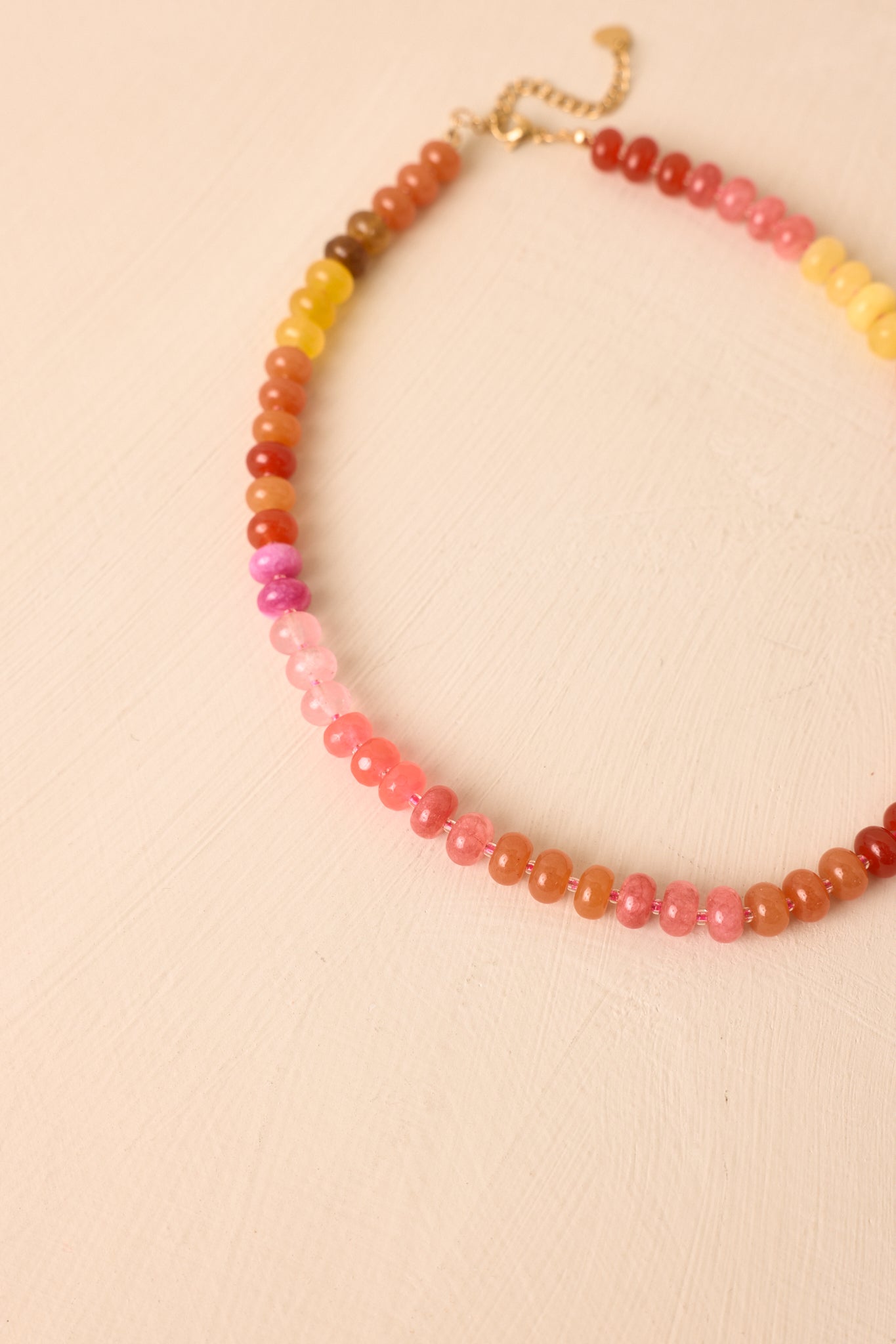 Colorful necklace arranged on a neutral beige background featuring mixed pink, coral, orange, and yellow beads with an adjustable chain extender.