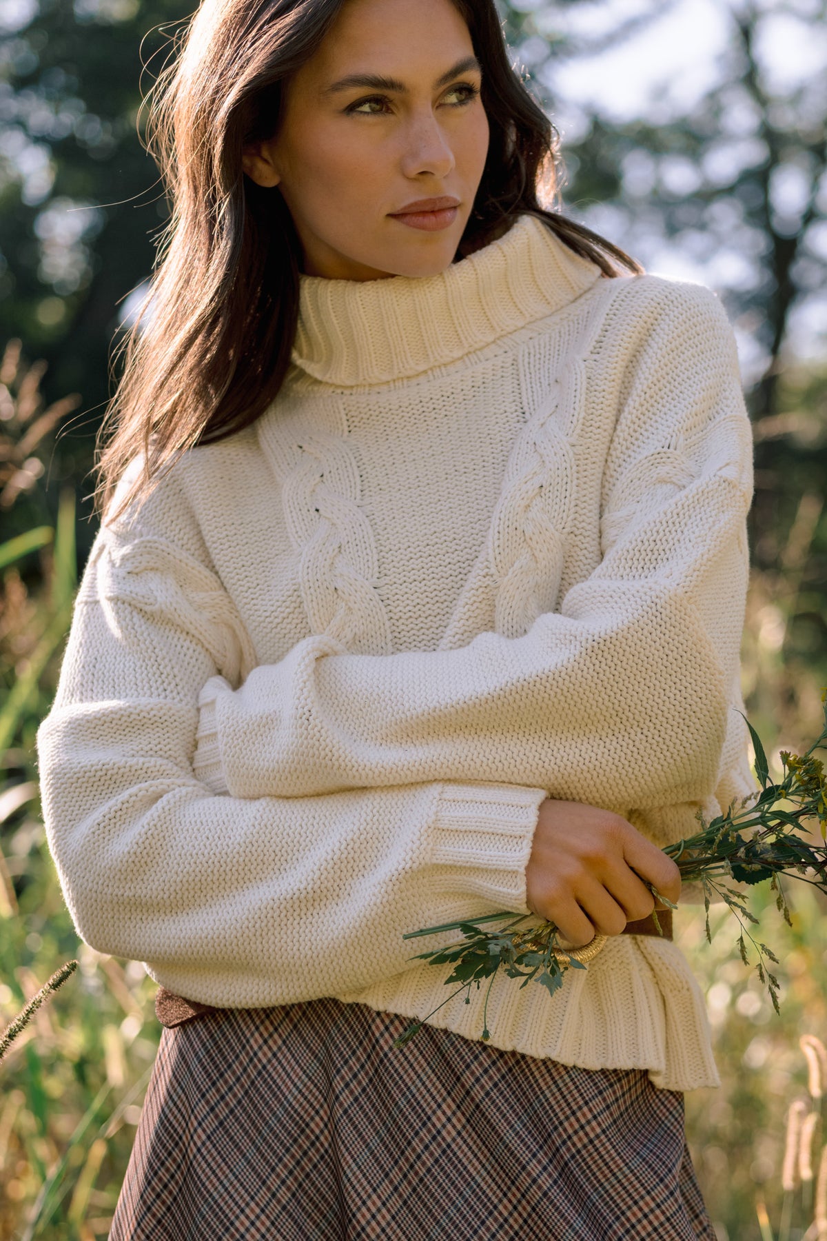 Model wearing a cream cable-knit turtleneck sweater with long sleeves and ribbed cuffs, styled with a plaid skirt.