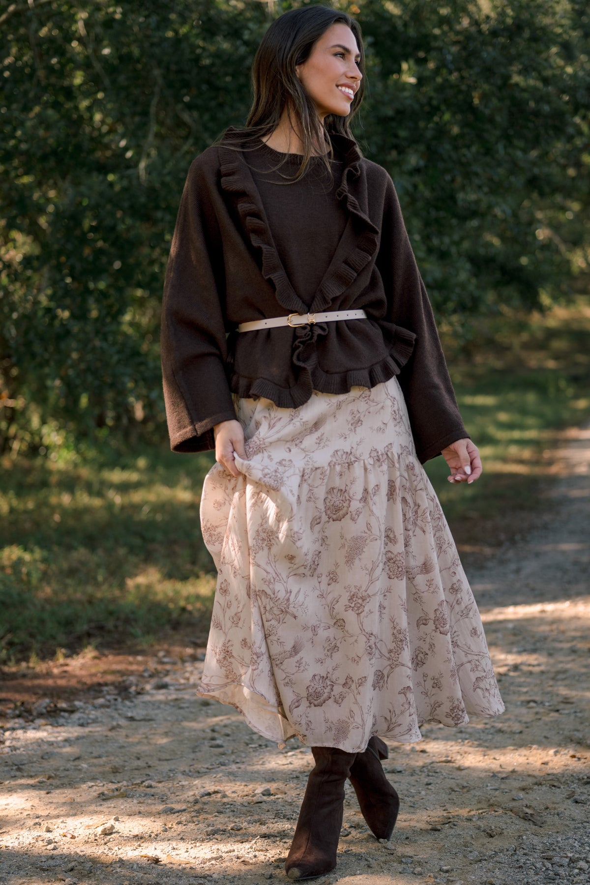 Model wearing a chocolate brown long-sleeve ruffle-trim top with a slim cream belt, styled with a cream floral midi dress and brown boots.