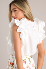 Angled back view of this ivory top that features a high crew neckline, a keyhole with a button closure at the back of the neck, and ruffled shoulder detailing.