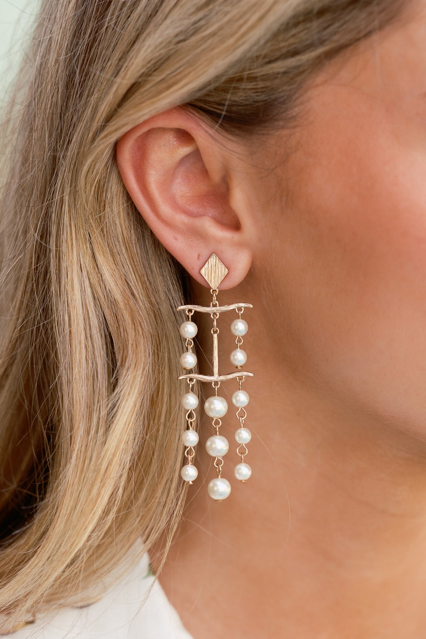 Cropped front view of the earrings showing the diamond-shaped gold stud, gold T-shaped drop charm, and hanging chains with ivory faux pearls.