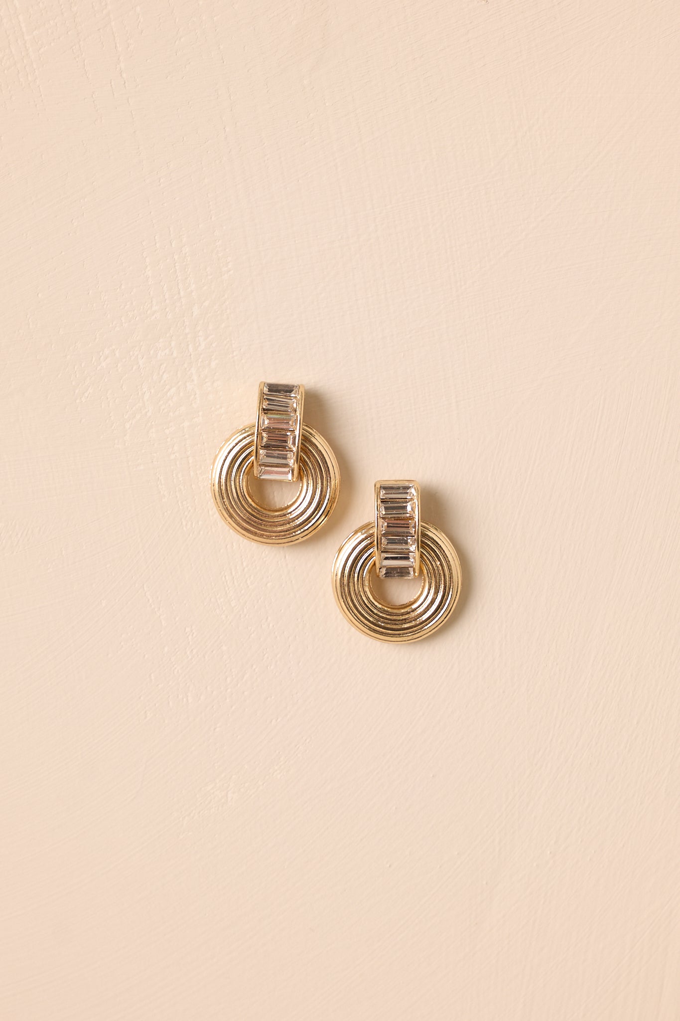 Gold earrings displayed against a beige background, showcasing the circular drop design and secure post backing.