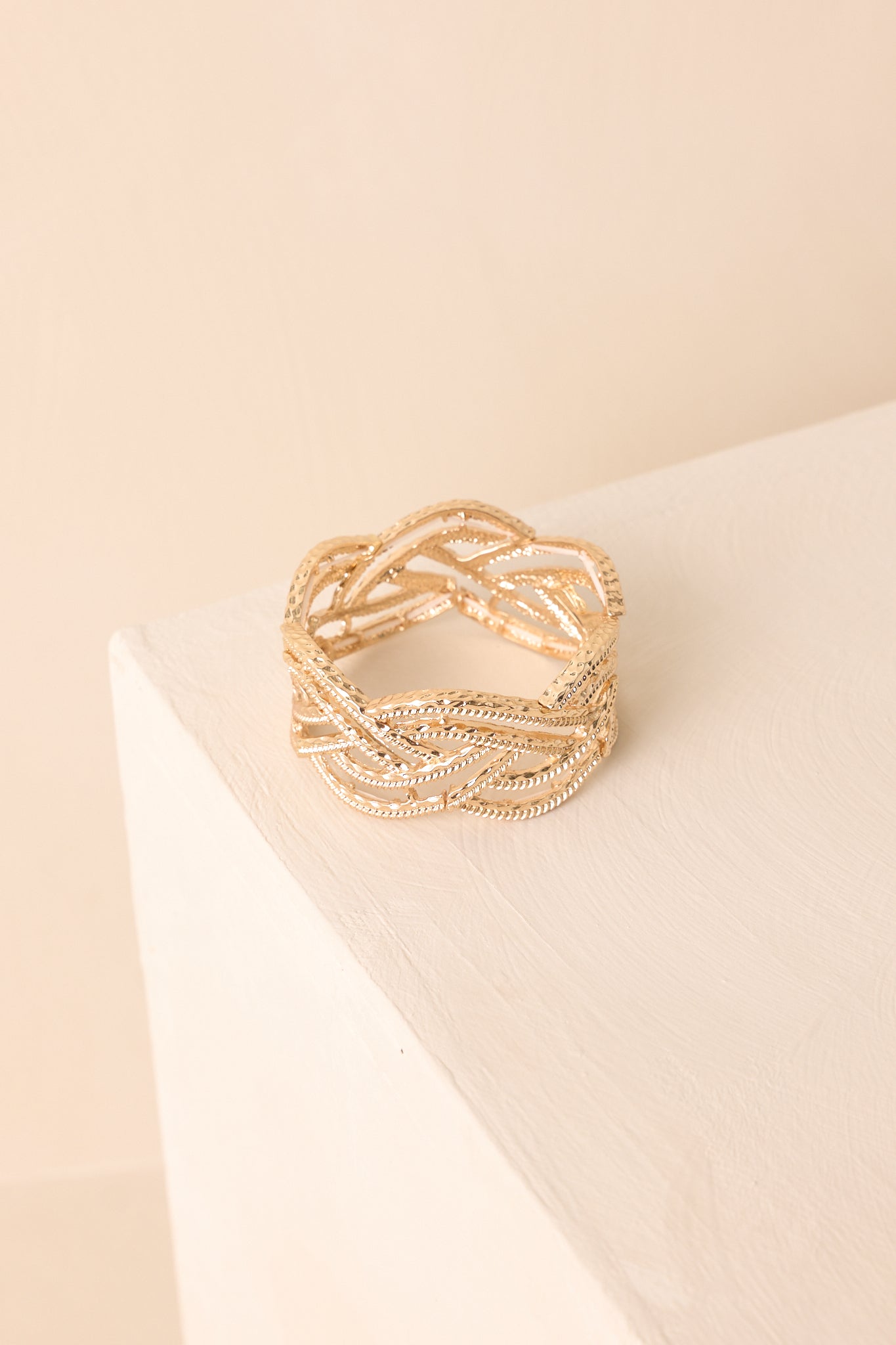 The slip-on gold bracelet featuring an open braided design and textured finish shown against a beige background.