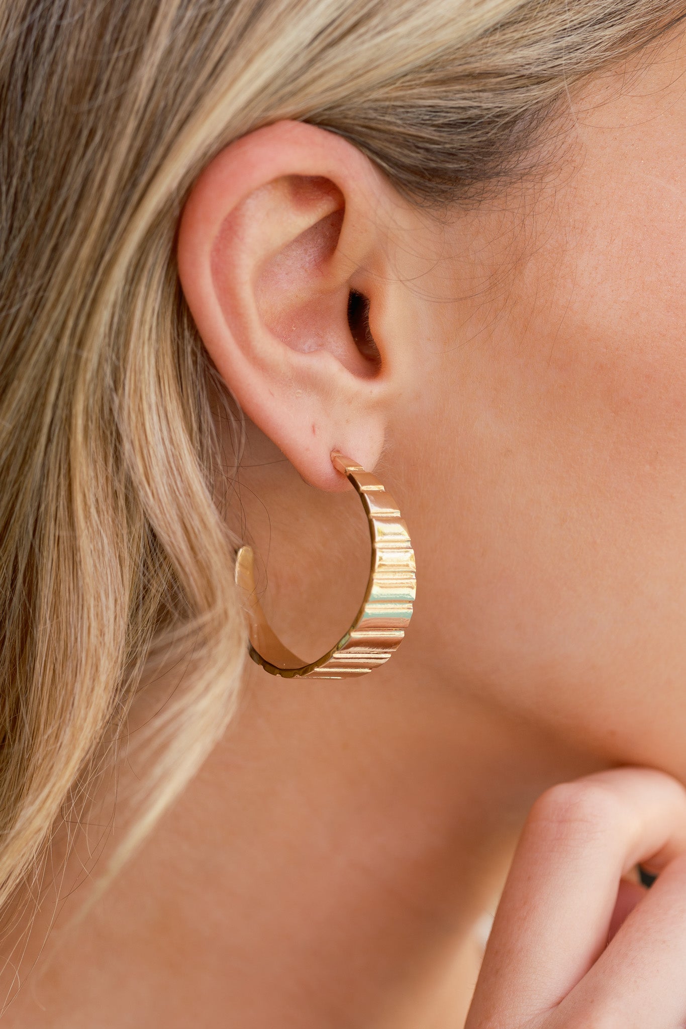 Gold earrings with an open hoop design, vertical ribbed texture, wide center, tapered C-hoop shape, and secure post backings, shown worn on a model’s ear.