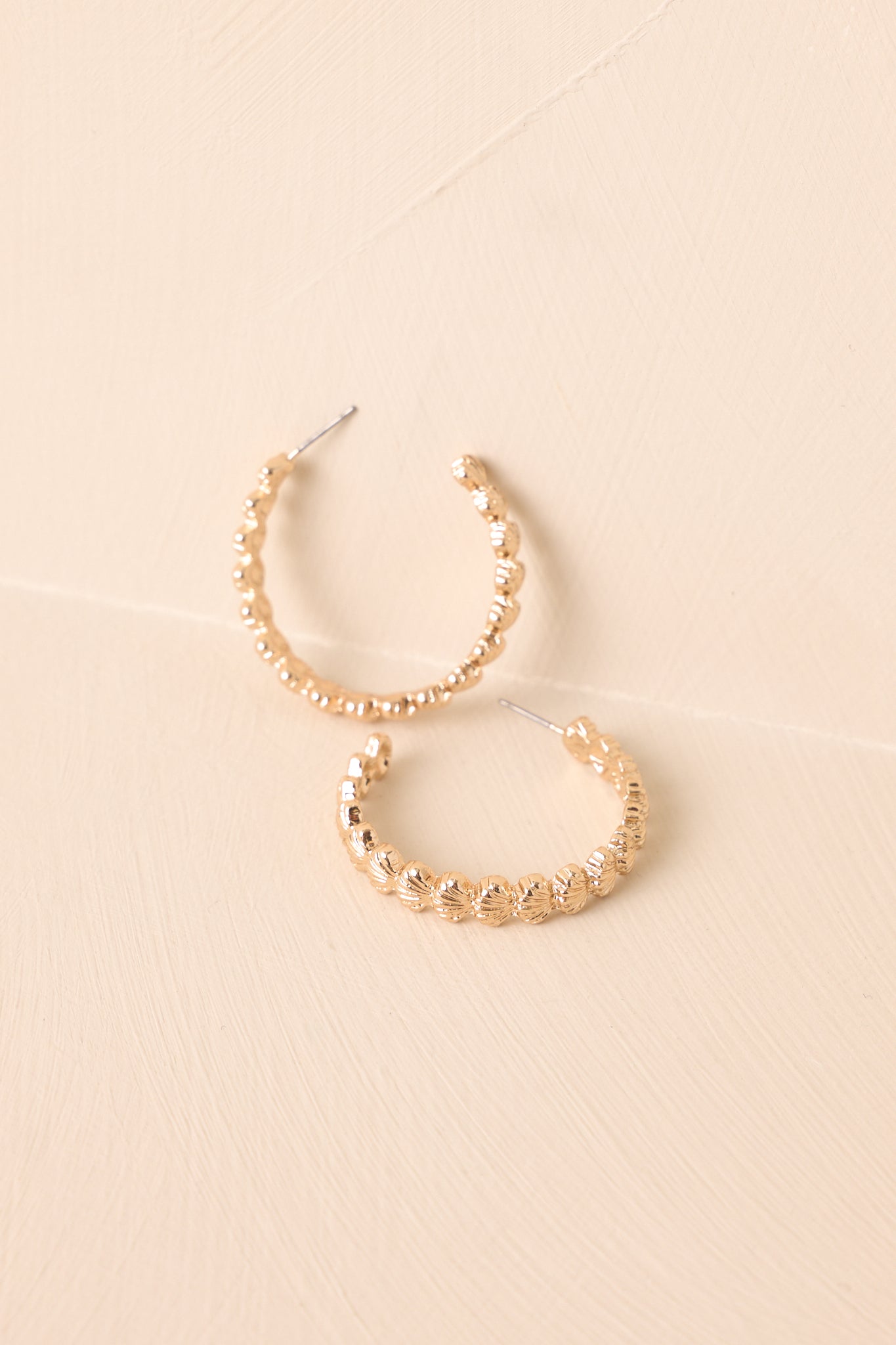 Single gold scalloped hoop earring with textured seashell edges propped up on neutral backdrop