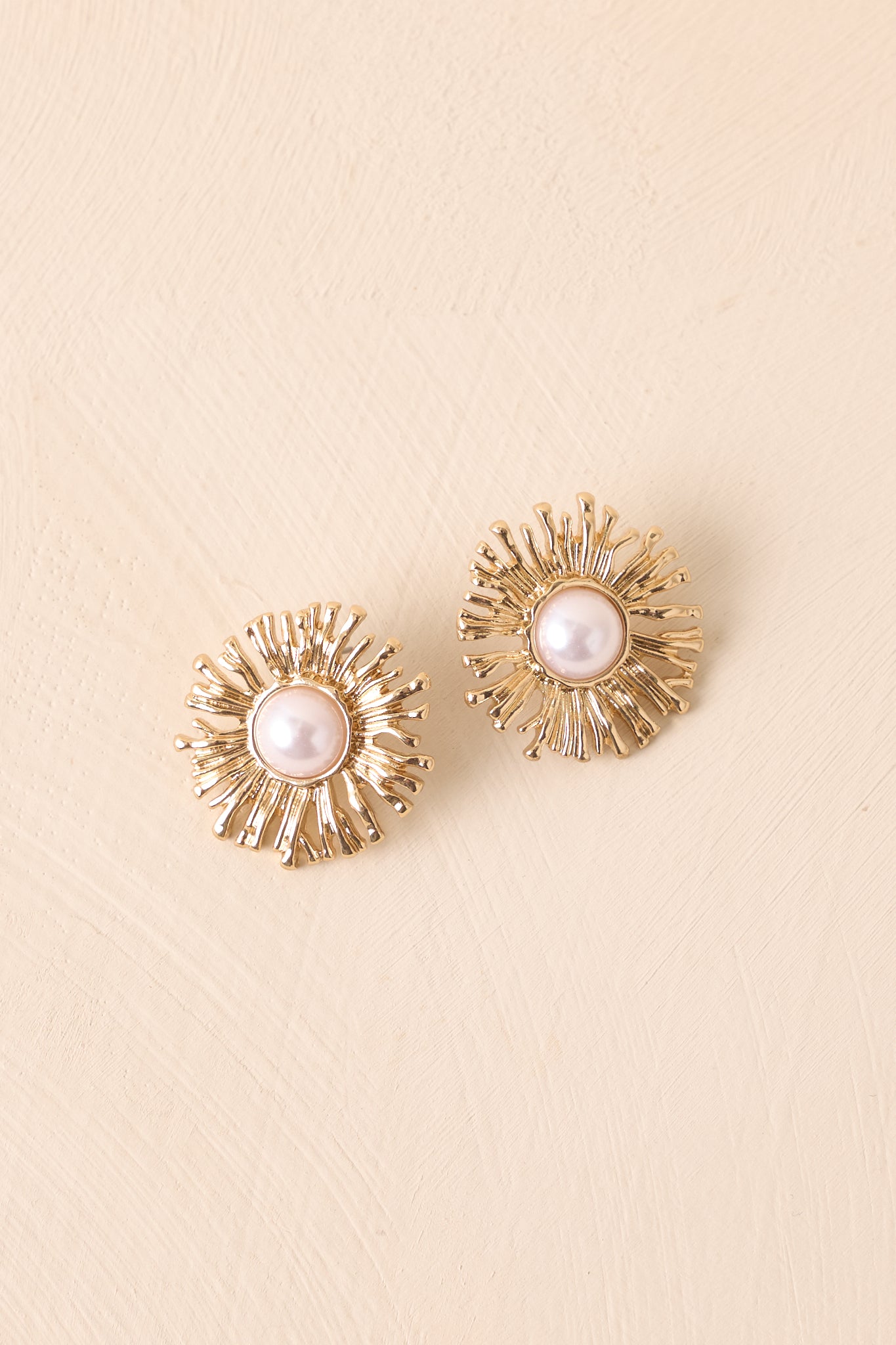 Gold sunflower stud earrings with textured petals and an ivory faux pearl center displayed on a neutral backdrop.