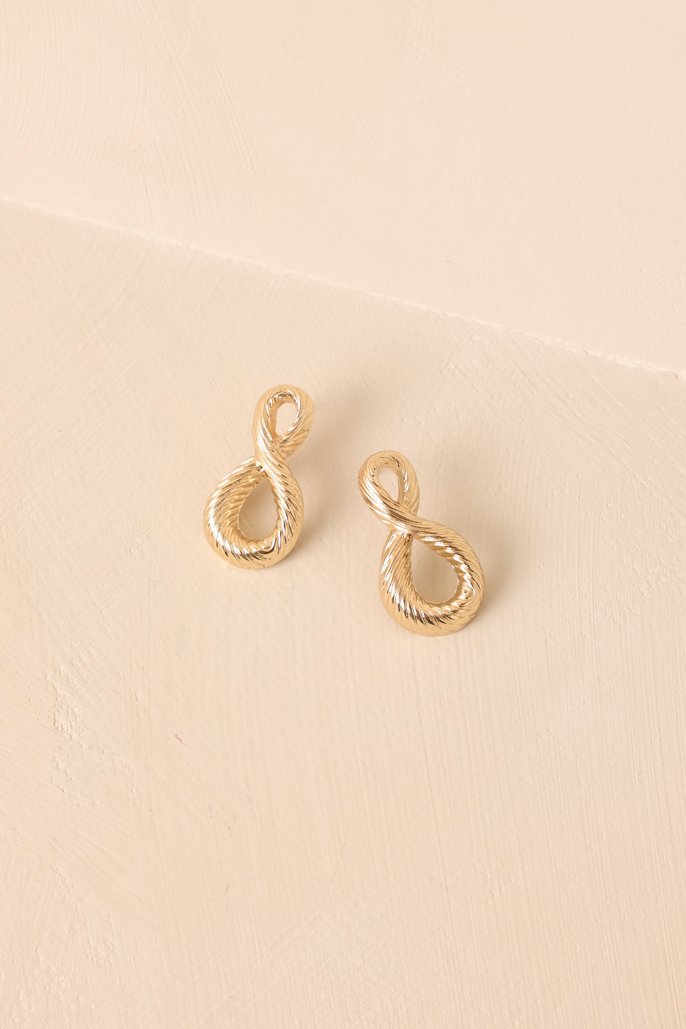 View of the gold-tone earrings highlighting the abstract teardrop silhouette and twisted rope design on beige background.