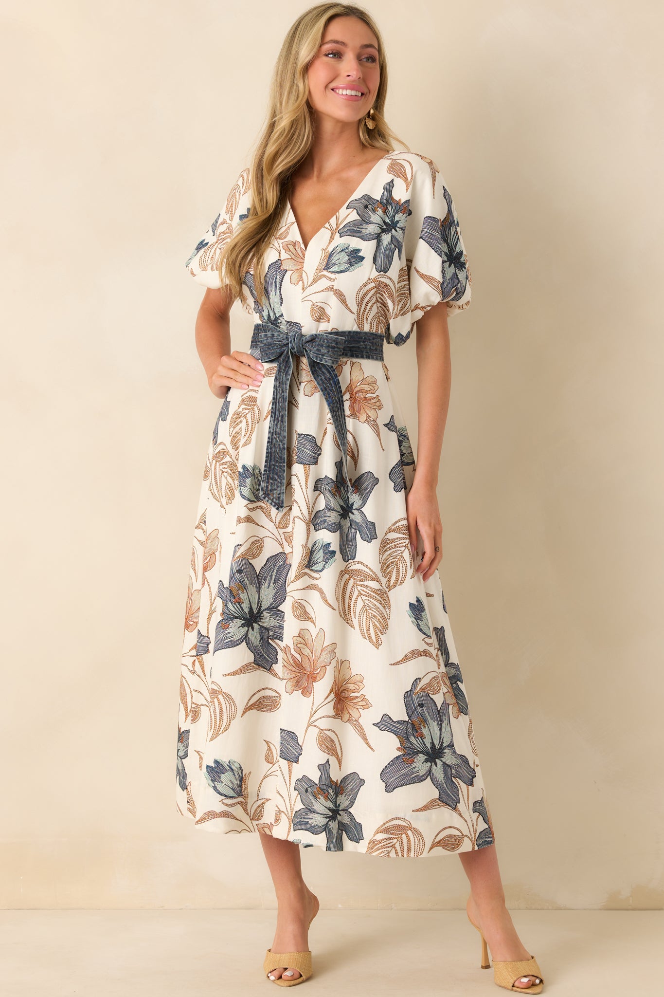 Woman wearing a cream midi dress with a blue floral print and wide puff sleeves, with a denim tie belt and woven heels.