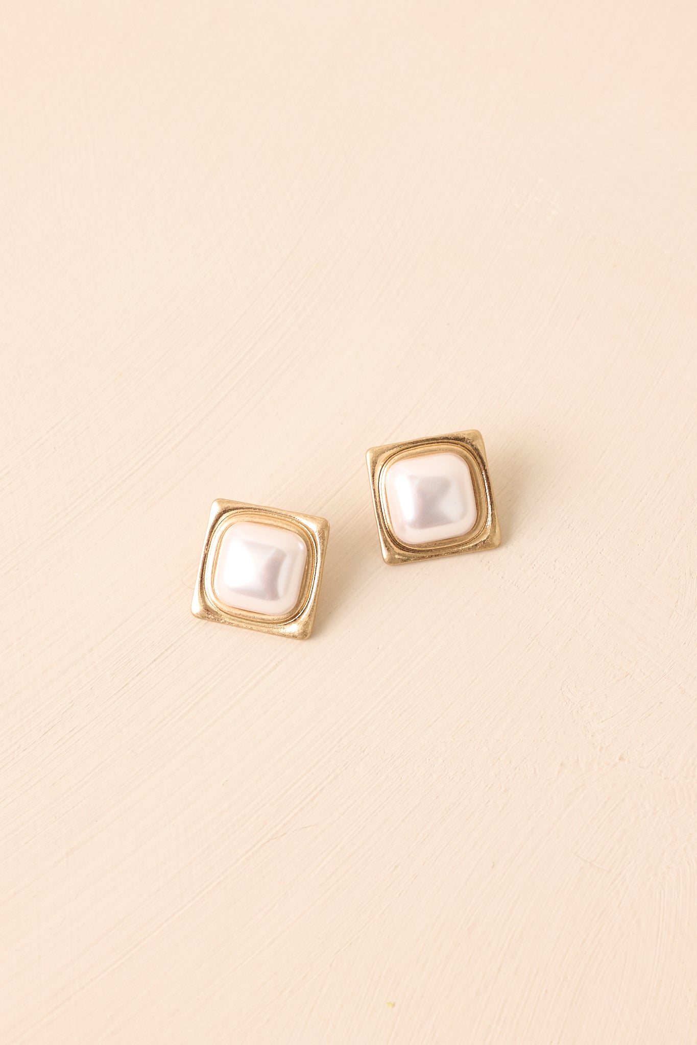Earrings highlighting the ivory faux pearl center and the textured square gold-tone frame on beige background.