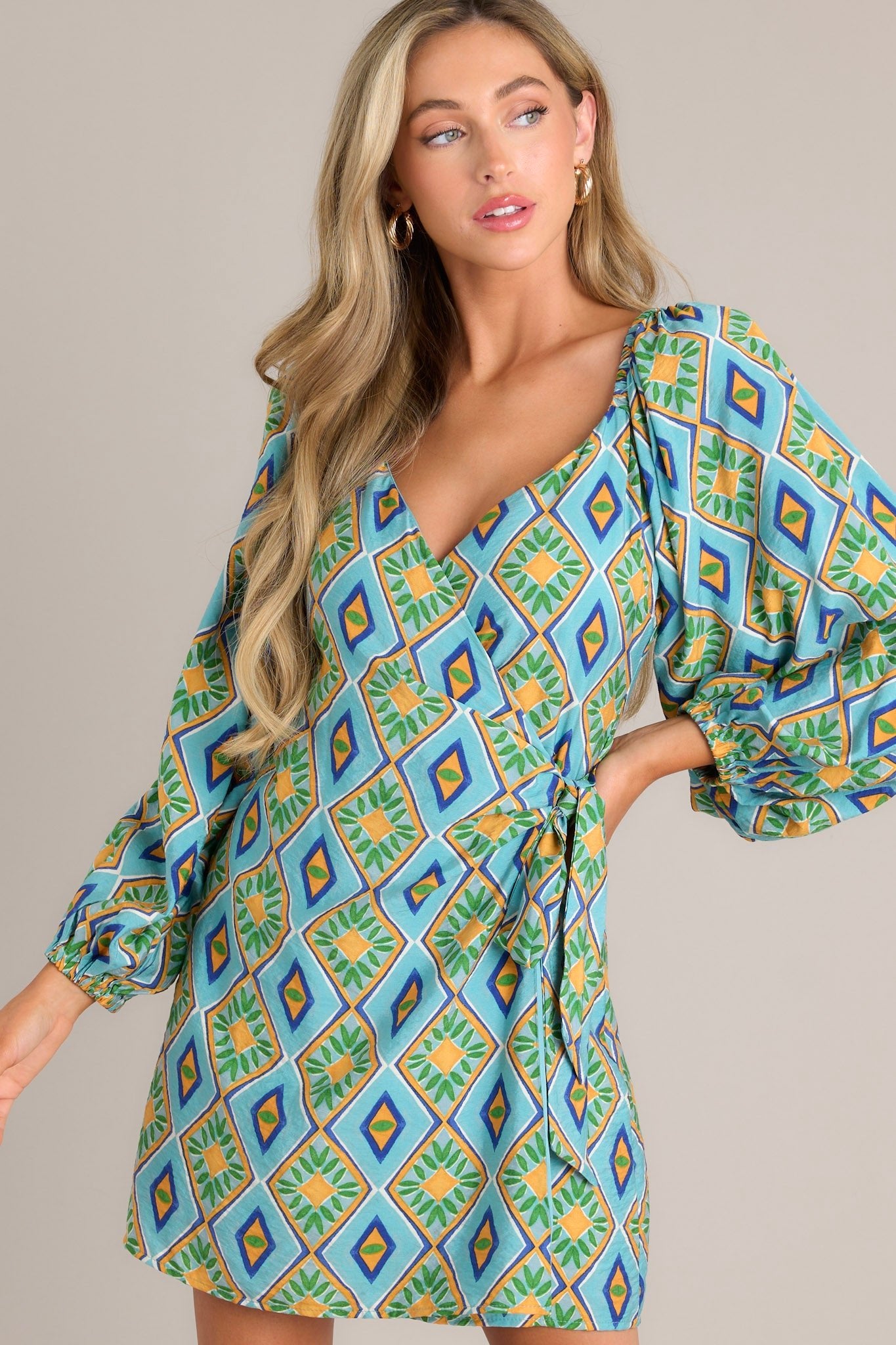 Angled front view of this turquoise multi print dress that features a wide v-neckline, a self-tie waist feature, elastic shoulders, and elastic cuffed sleeves.