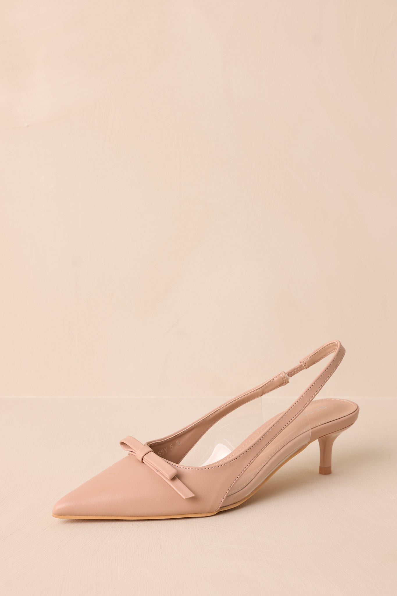 Front angled view of beige kitten heels featuring a pointed closed toe, delicate bow detailing, a strap around the back of the foot, clear siding for extra support, and an extremely short heel