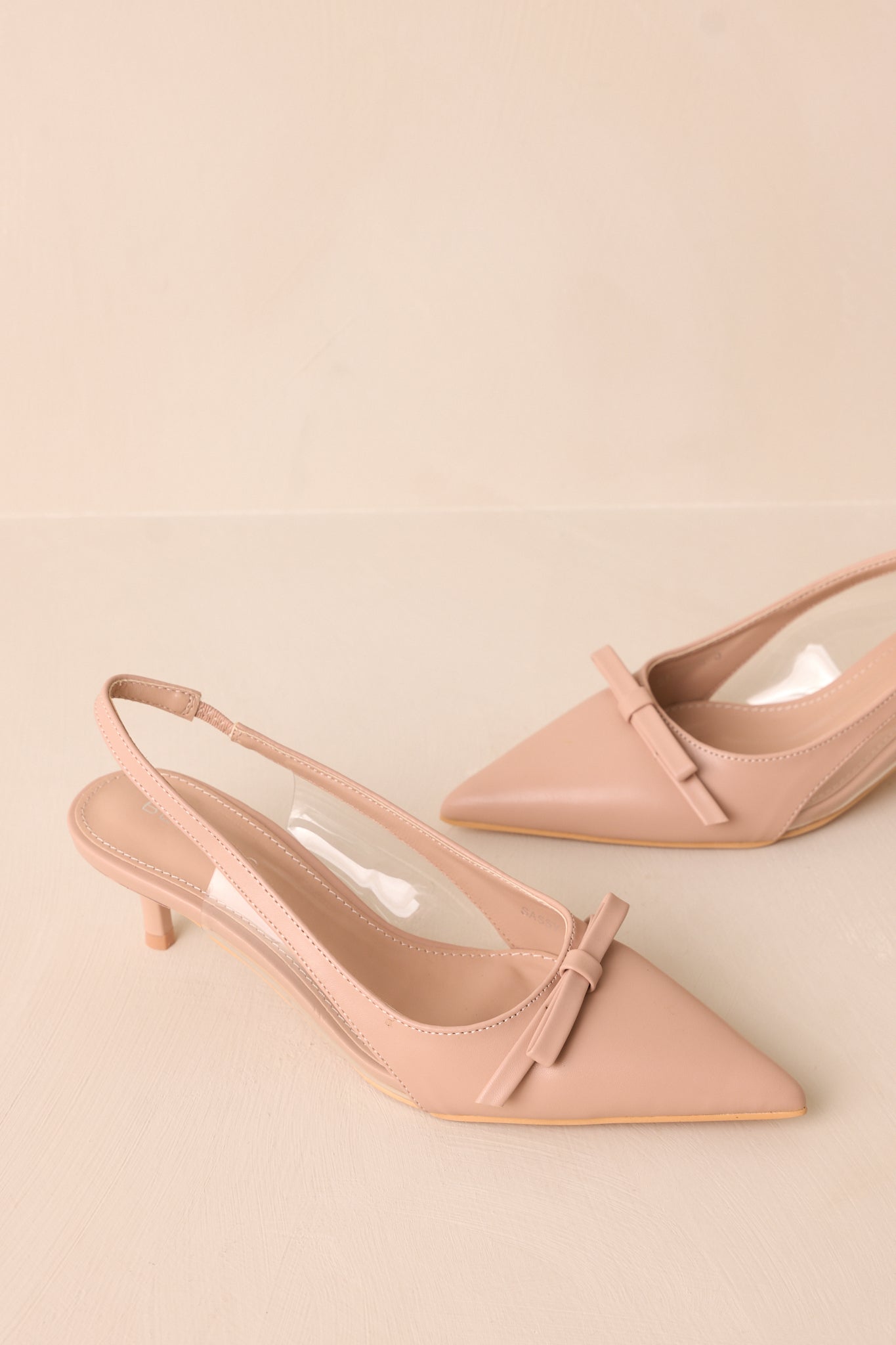 Front angled view of beige kitten heels featuring a pointed closed toe, delicate bow detailing, a strap around the back of the foot, clear siding for extra support, and an extremely short heel