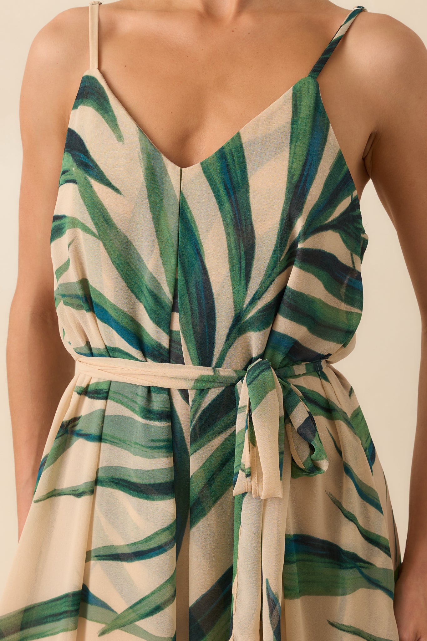 Close-up detail photo of model wearing a beige maxi dress that features a green palm print chiffon overlay, beige lining, deep v-neckline, adjustable spaghetti straps, and an optional self-tie belt.