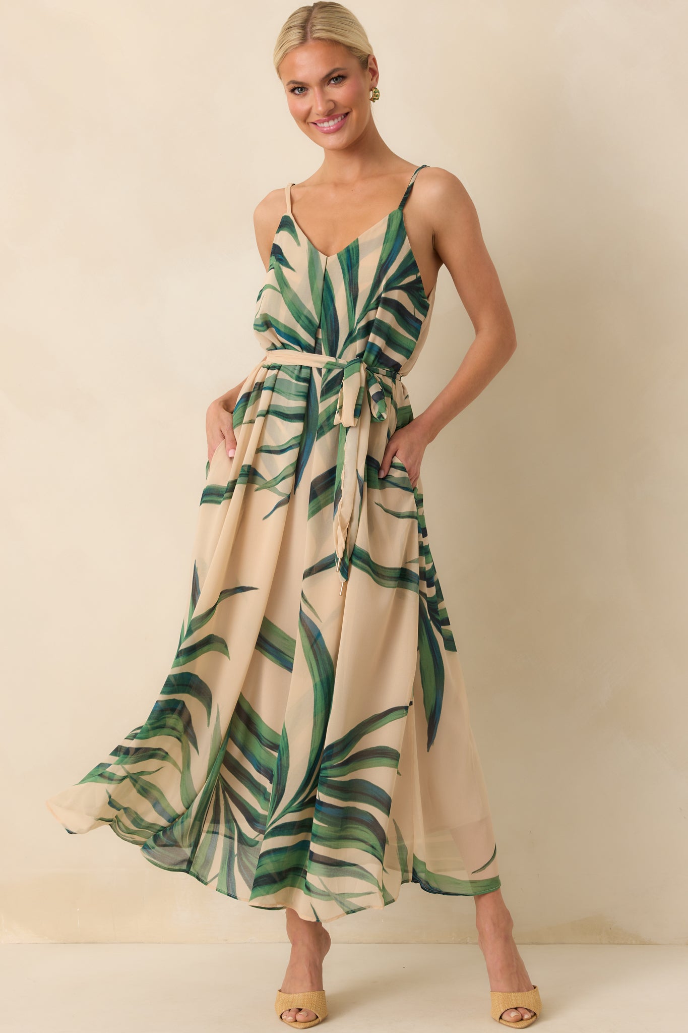 Full body of model wearing a beige maxi dress that features a green palm print chiffon overlay, beige lining, deep v-neckline, adjustable spaghetti straps, and an optional self-tie belt.
