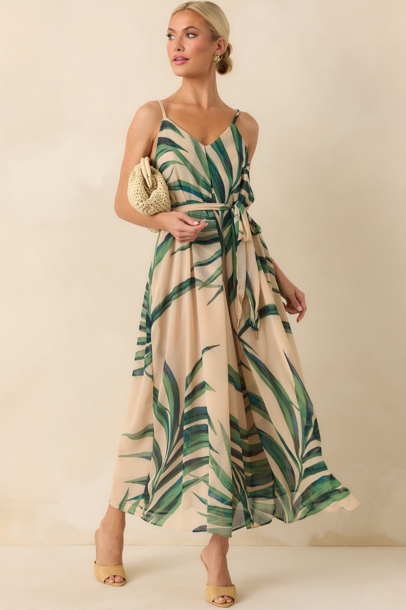 Full body photo of model walking in a beige maxi dress that features a green palm print chiffon overlay, beige lining, deep v-neckline, adjustable spaghetti straps, and an optional self-tie belt.
