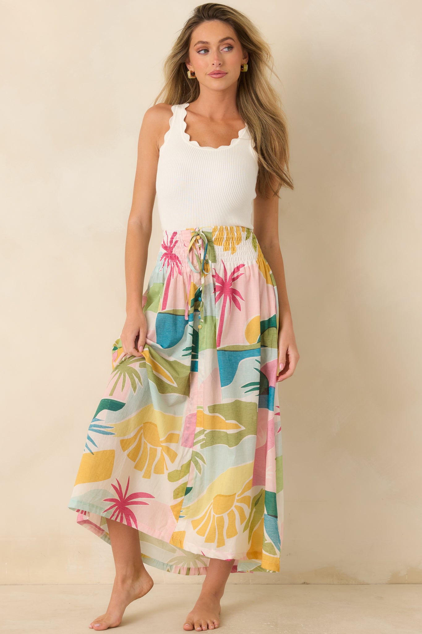 Model wearing the Paradisa flowy tropical print maxi skirt with a smocked elastic waistband, adjustable drawstring tie, side pockets, and colorful pastel tropical landscape print.