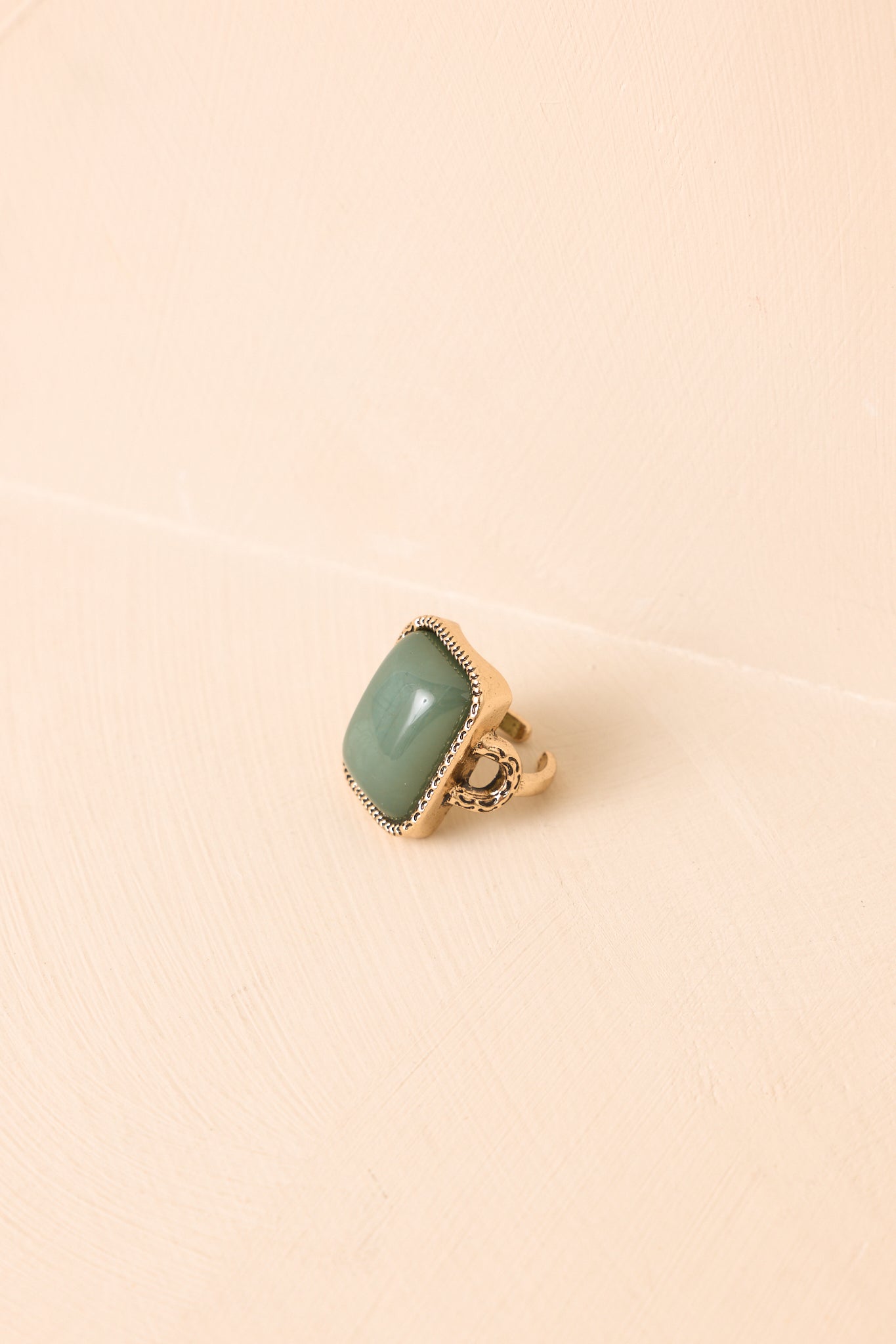 Rectangular sage green stone statement ring with an antique gold adjustable band displayed on a neutral textured background.