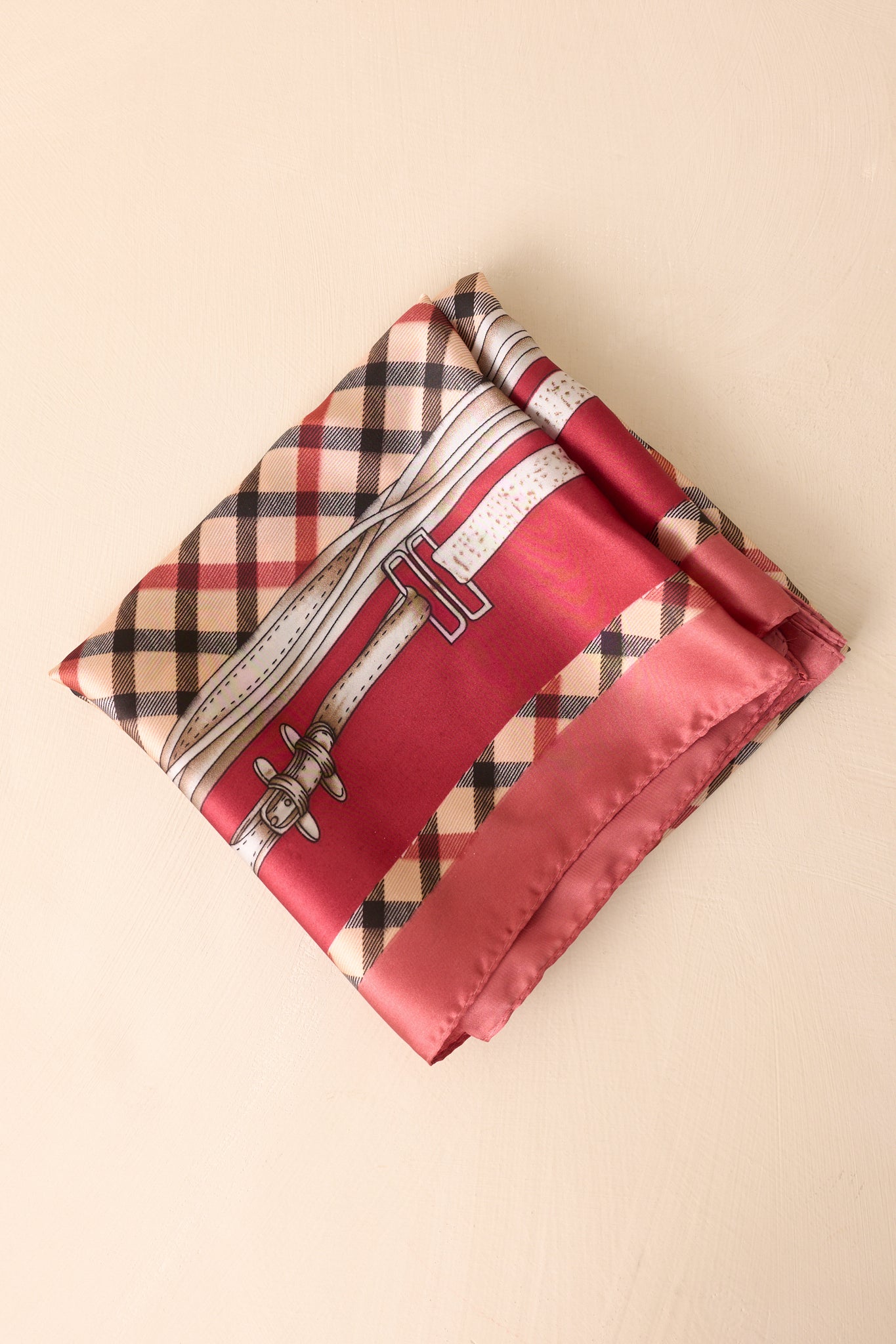 Square scarf neatly folded into a smaller square, displaying the burgundy border framing the classic beige, black, and red plaid pattern with hints of the equestrian chain and buckle motifs.