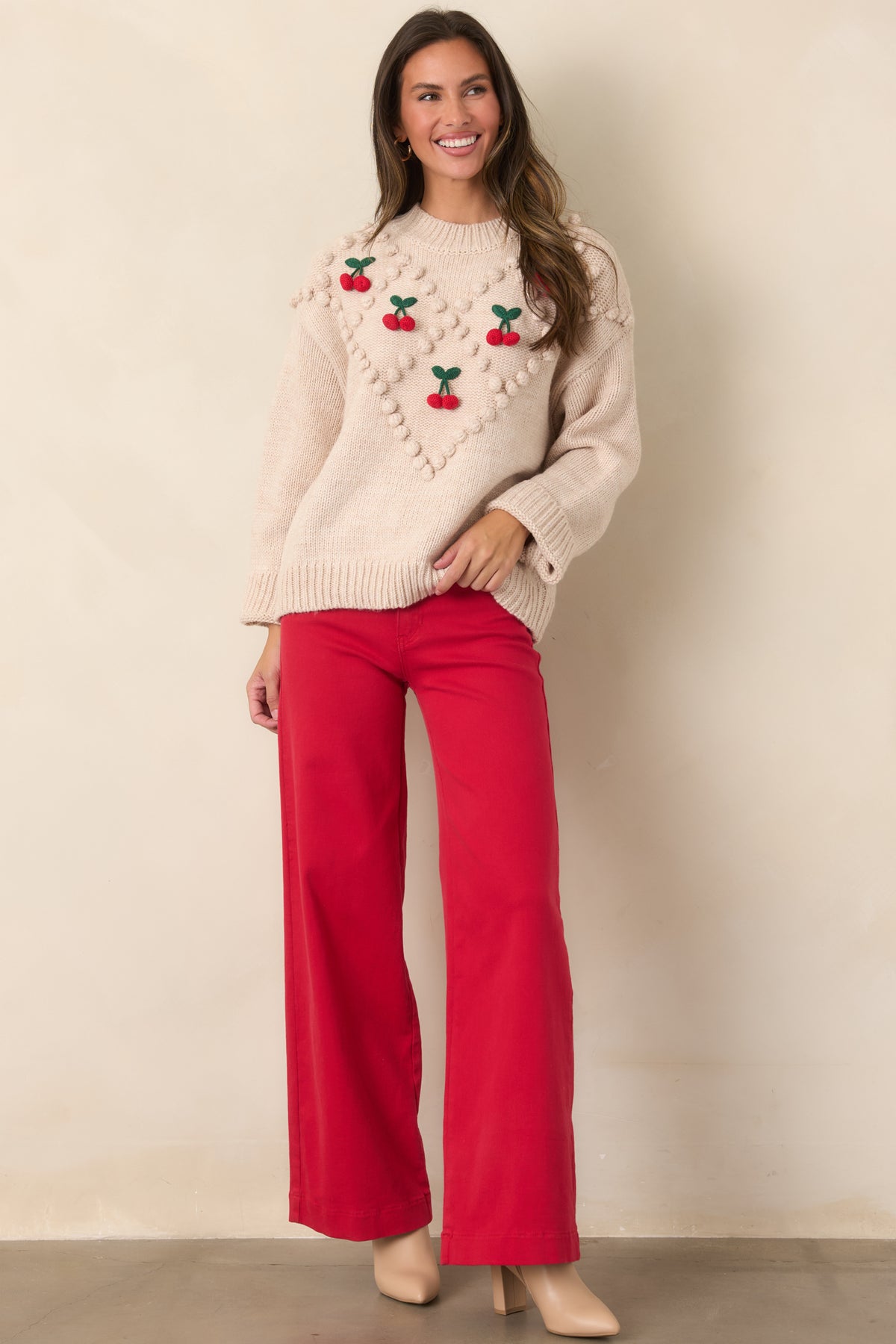Model wearing a relaxed beige knit sweater with red cherry accents, green leaves, and diamond-pattern bobble detailing.