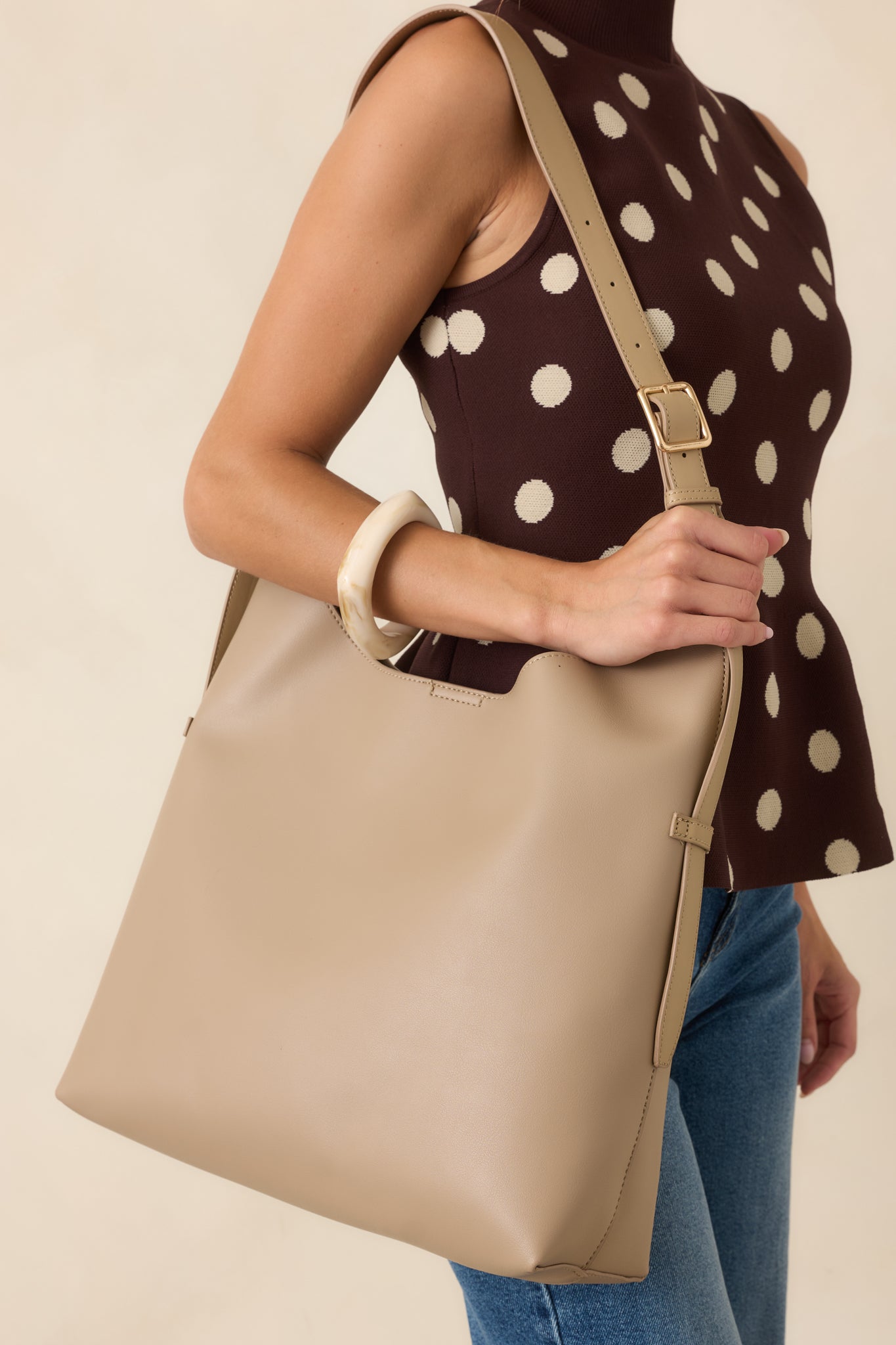 Close-up of a taupe slouchy shoulder tote bag with an adjustable strap, styled with a brown sleeveless polka dot mock-neck top, blue jeans, and a cream resin bangle.