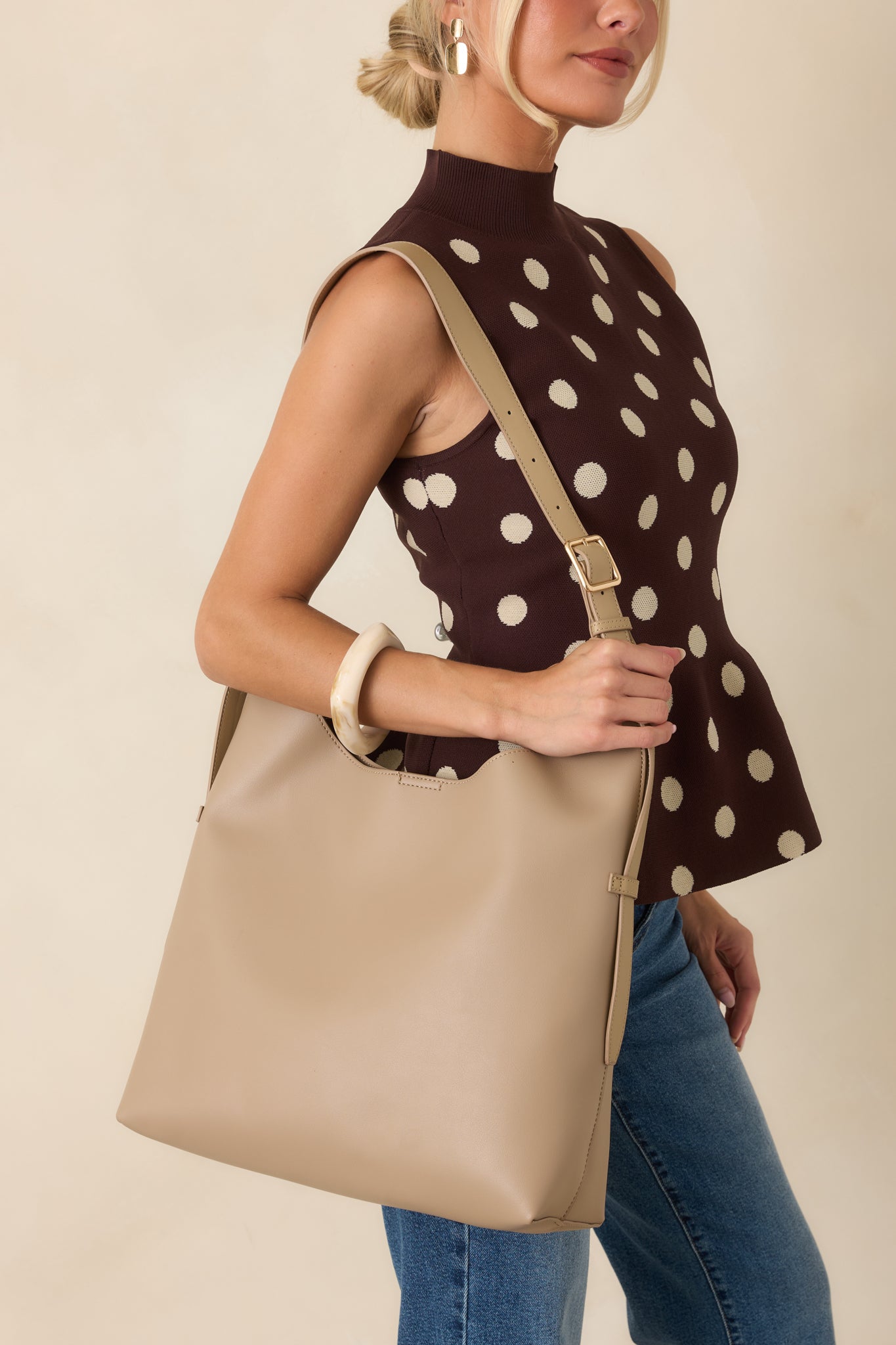 Taupe slouchy shoulder tote bag with an adjustable strap worn over the shoulder, styled with a brown polka dot mock-neck sleeveless top, blue jeans, gold earrings, and a cream resin bangle.