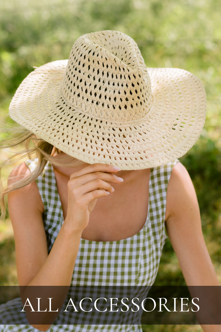 Model wearing a woven straw sun hat and green gingham dress while sitting outdoors in soft natural light. Hat featured in All Accessories shop.