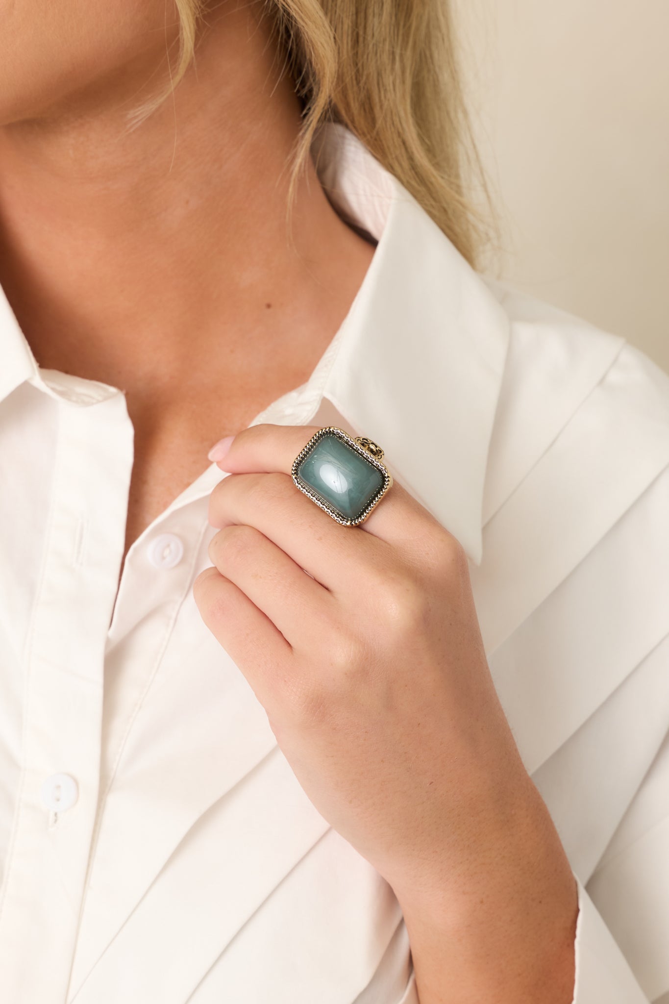 Sage green statement ring with an antique gold setting styled on a woman's hand with a white blouse for an elegant everyday look.