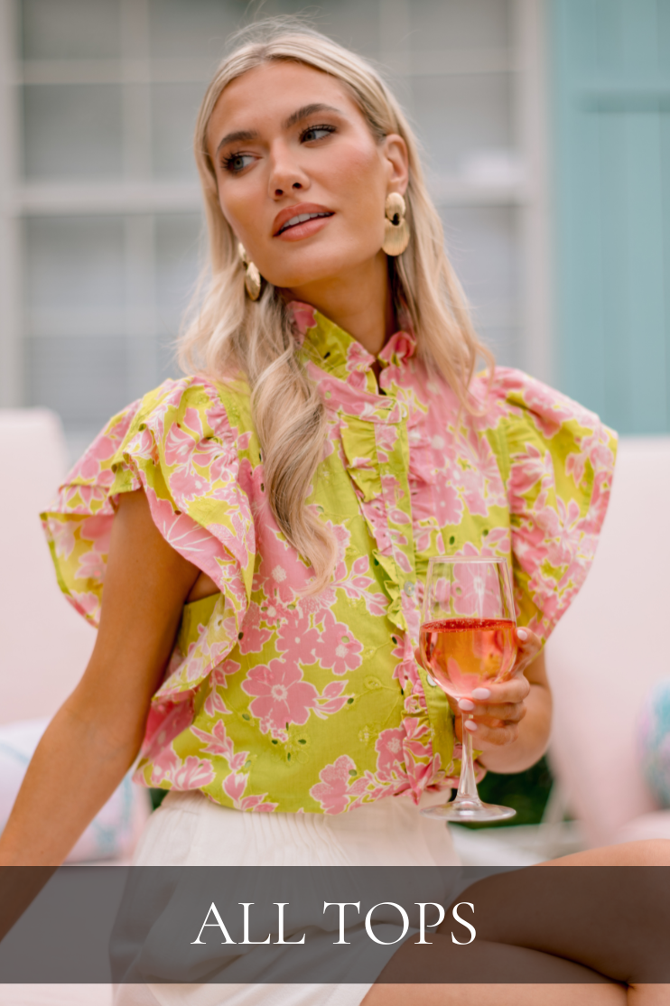 Model wearing a pink and lime floral ruffle blouse with statement earrings, holding a glass while seated outdoors in a resort setting. Top is featured in All Tops.