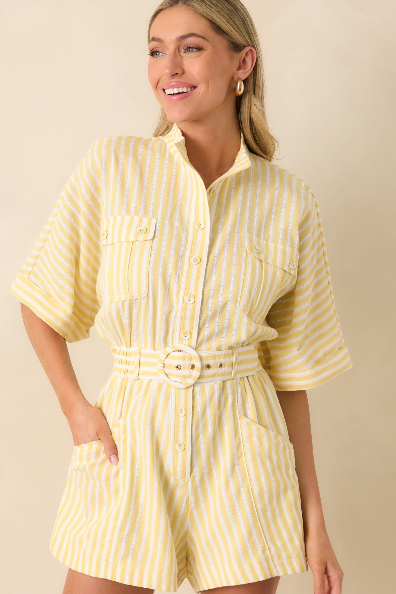 Close-up of yellow striped romper featuring detailed stitching, collared neckline, button front, cuffed short sleeves, belted waist, and front patch pockets.