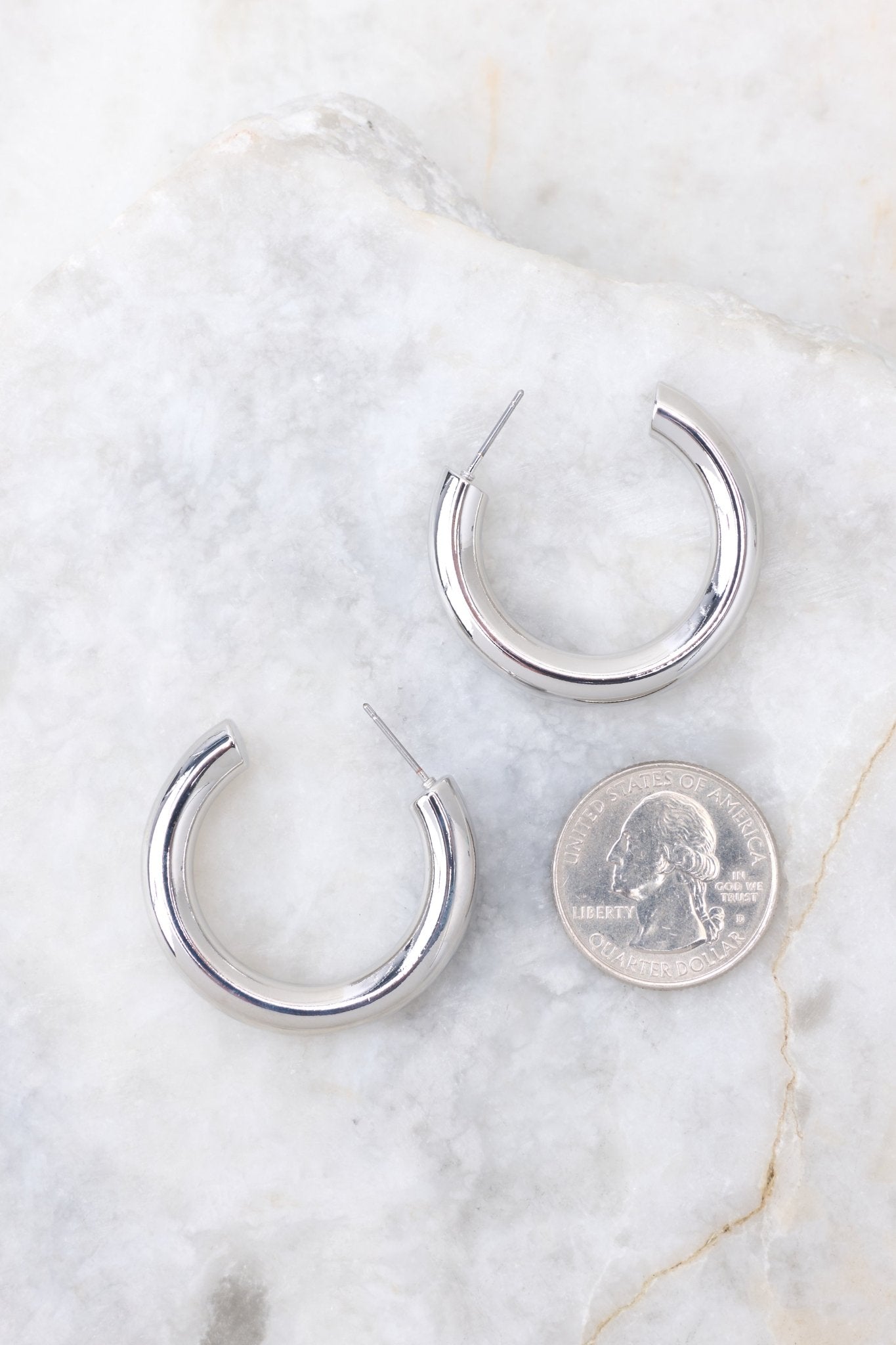Silver hoop earrings that feature a reflective finish compared to quarter to actual size. Earrings measure 1.5" in diameter.