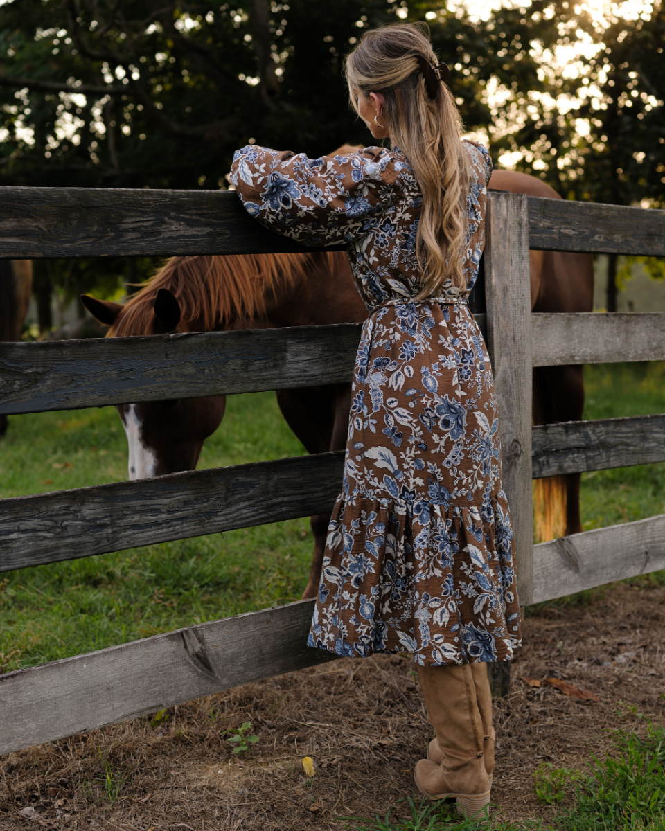 Woman in a brown midi dress with a blue paisley print, paired with boots and standing in front of a fence with horses behind it. 