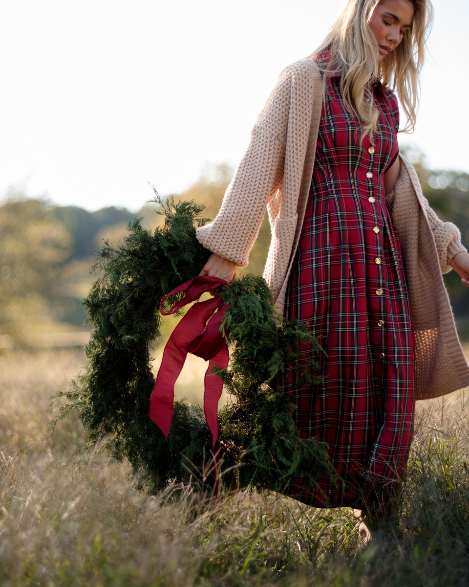 A red tartan plaid maxi shirt dress with a camel cardigan, carrying an evergreen wreath with a red ribbon