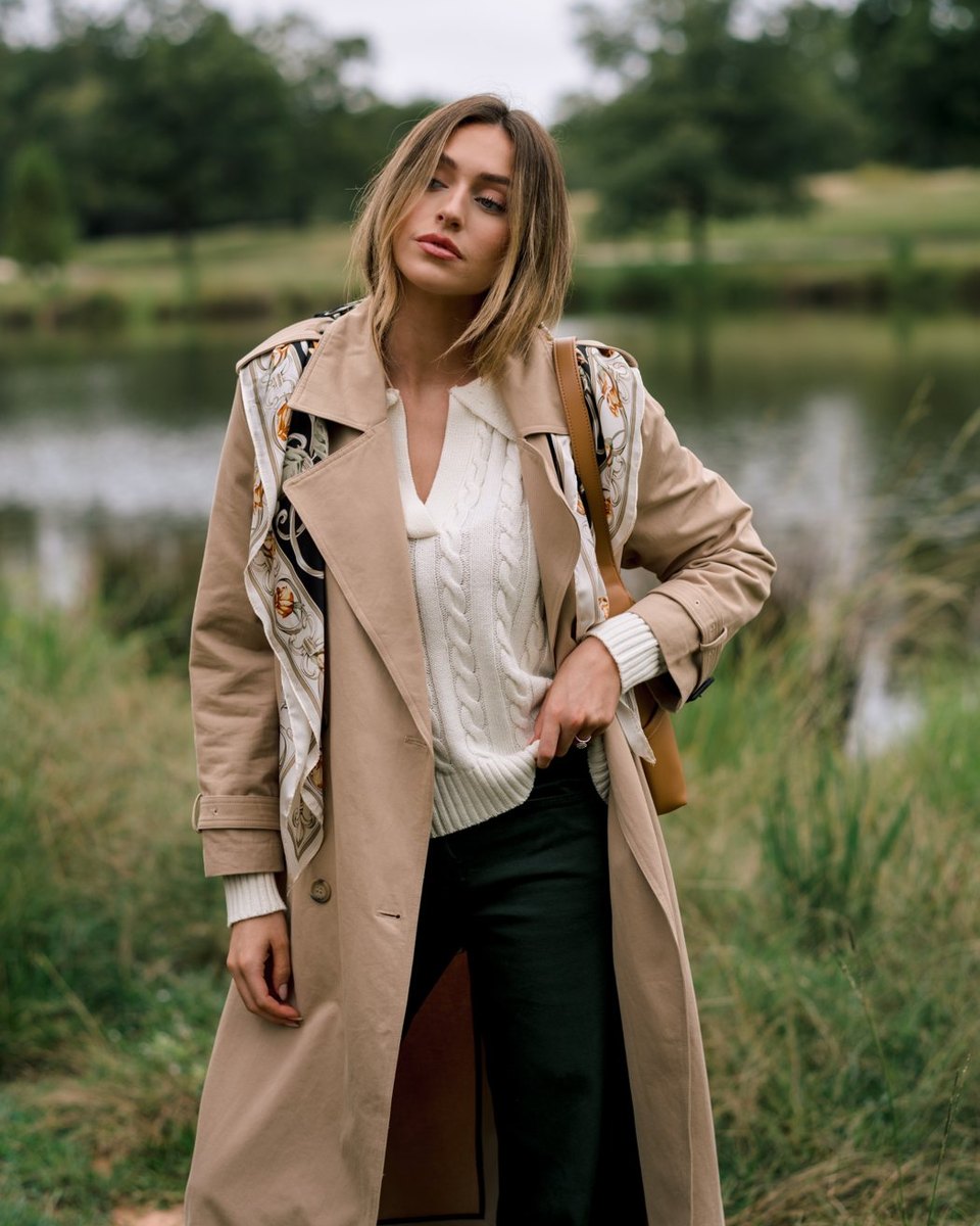 Woman wearing an open tan trench coat over an ivory cable knit sweater and dark trousers beside a pond.