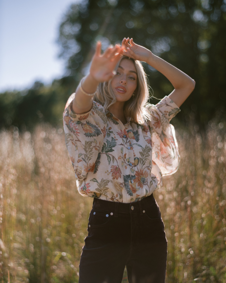Image of model out in a field wearing a floral blouse and brown jeans.