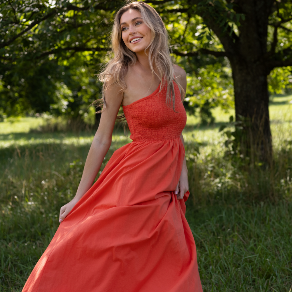 Woman wearing a coral one-shoulder smocked maxi dress that is featured in the Destination Vacation drop.