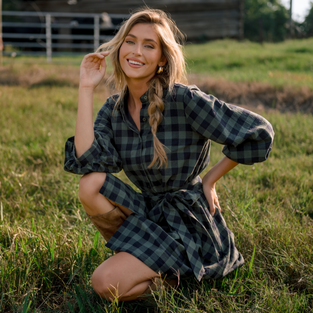 Woman wearing a dark green and black plaid dress, kneeling in a grassy field outdoors during golden-hour light, showcasing a cozy fall look.