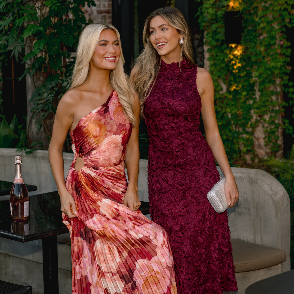 Two models standing outside by a white building wearing new arrival dresses, including a light blue mini dress and a blue, ivory, and orange floral maxi dress.