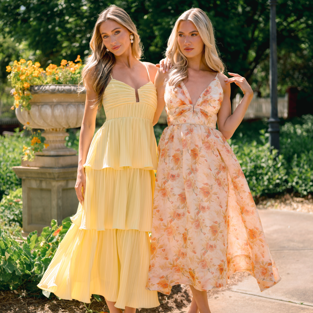 Two women in a garden wearing wedding guest dresses, including a yellow ruffle maxi dress and an orange floral maxi dress.