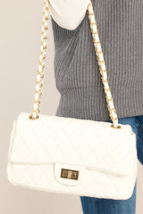 This white hang bag features a single flap, a chain strap, a turn lock closure, a hidden pocket, a zippered pocket on the inside, and gold hardware. 