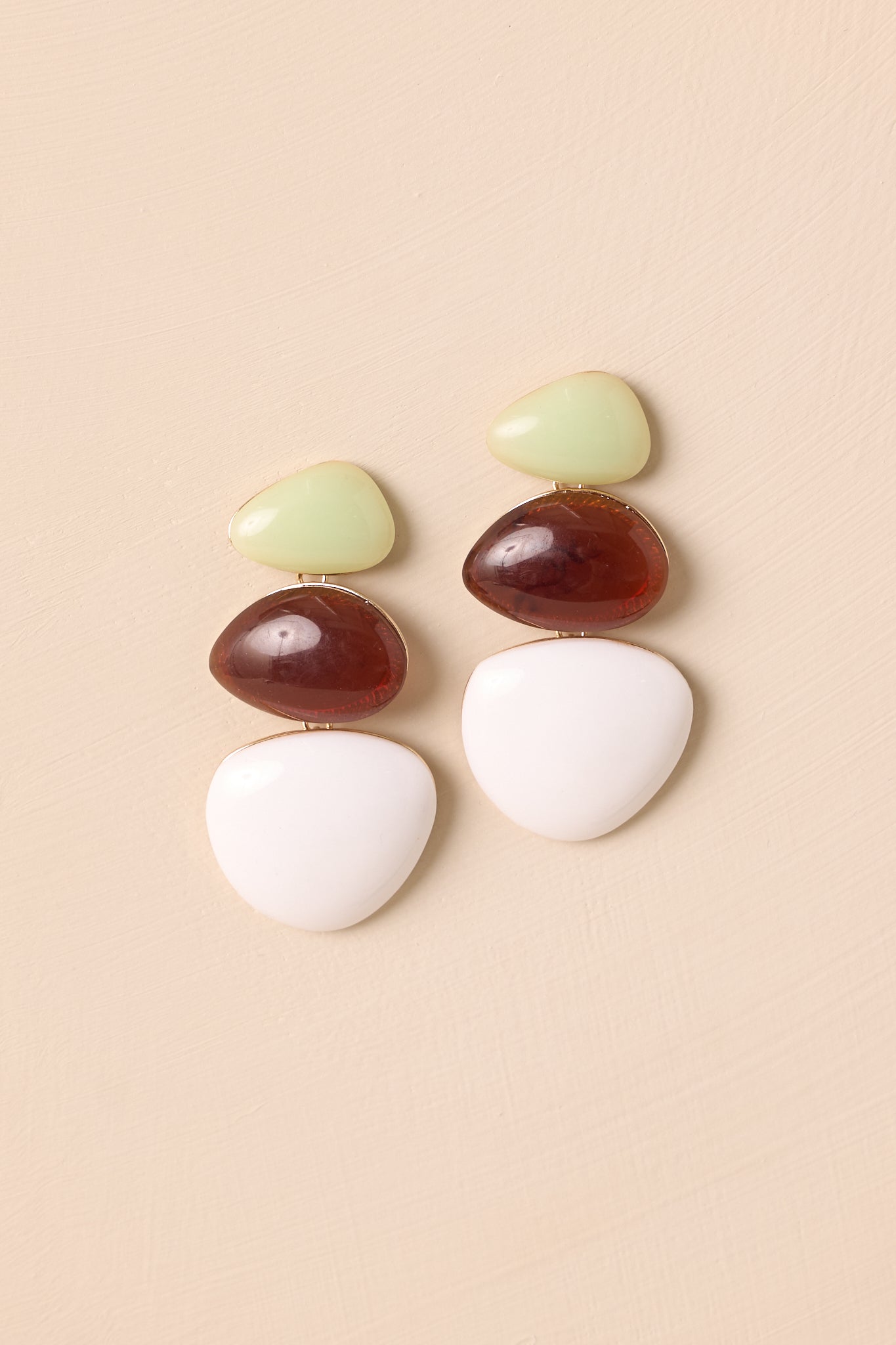 Earrings feature light green, brown, and white organic-shaped stones horizontally stacked in increasing size, set in a gold bezel with secure post backings on beige backdrop.