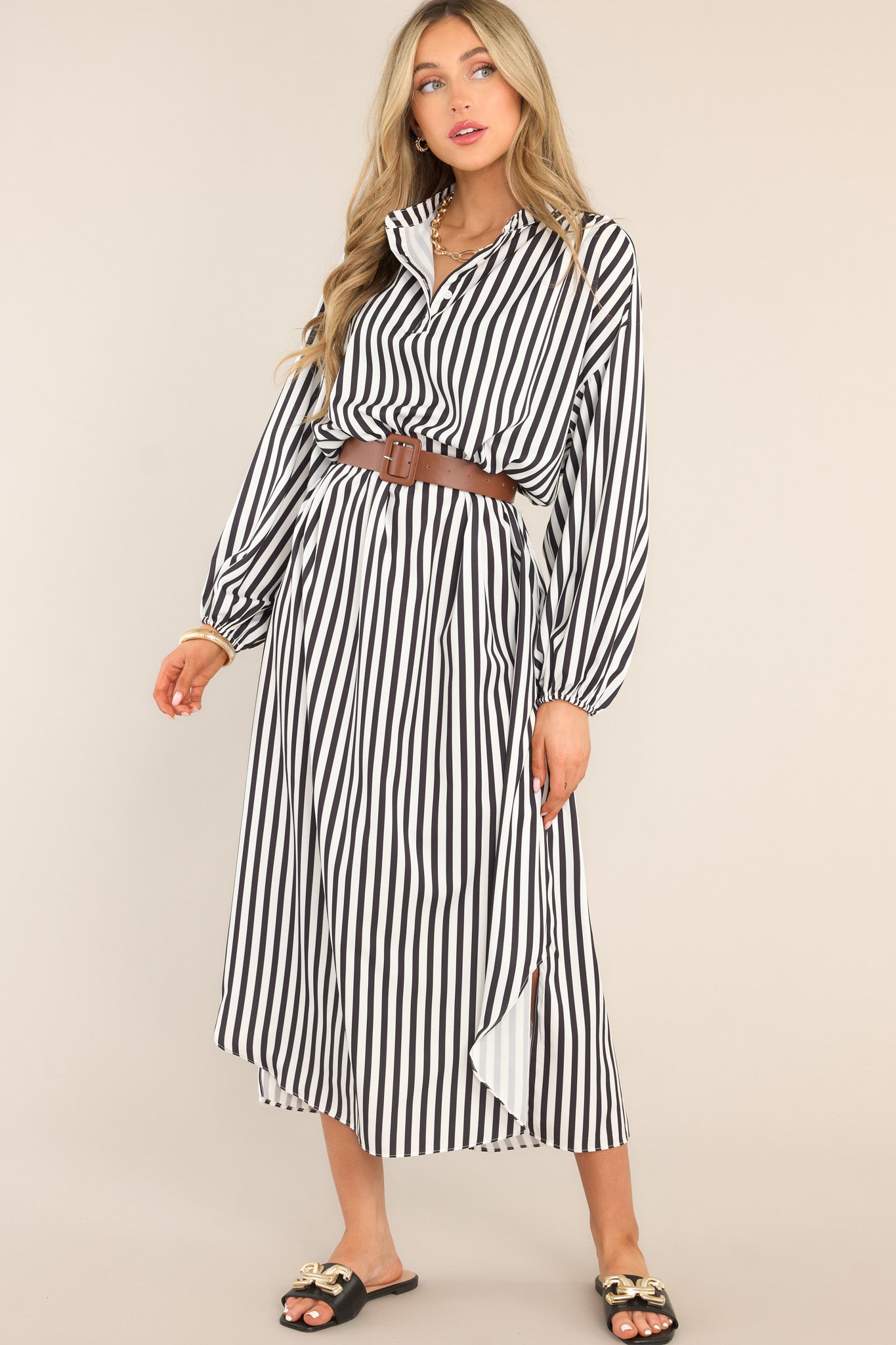 This black and white dress features a crew neckline, dropped shoulders, functional buttons, hip pockets, a functional belt, elastic cuffed long sleeves, and a split hemline.