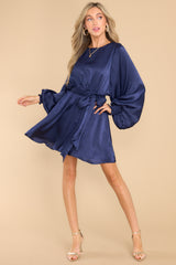 This navy blue dress features a round neckline, a keyhole with a button closure on the back, flowy sleeves with smocked cuffs, an elastic waistband, a self-tie, and a flowy skirt.