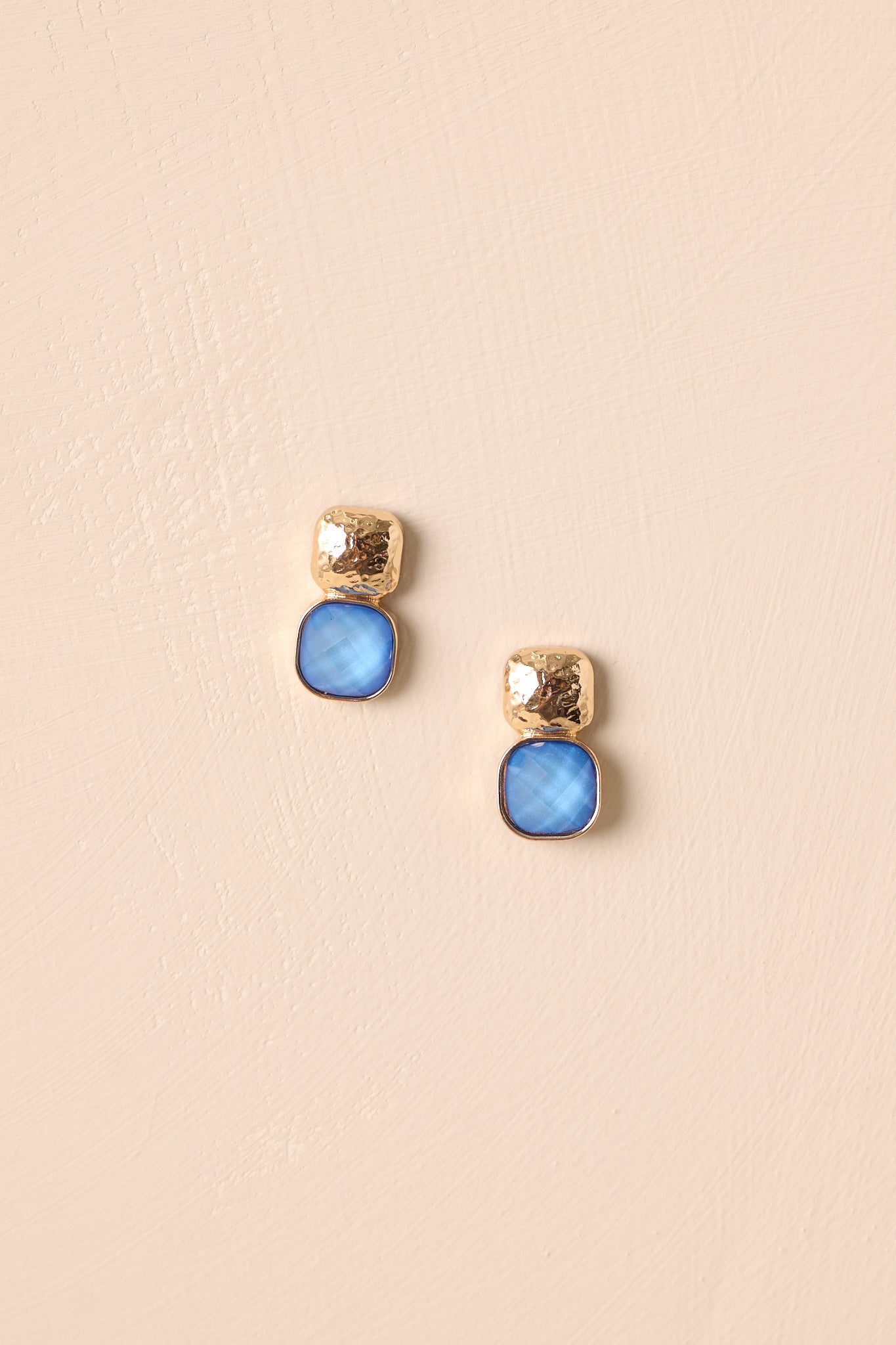 The blue cushion cut stone drop earrings with a hammered gold base shown against a beige background.