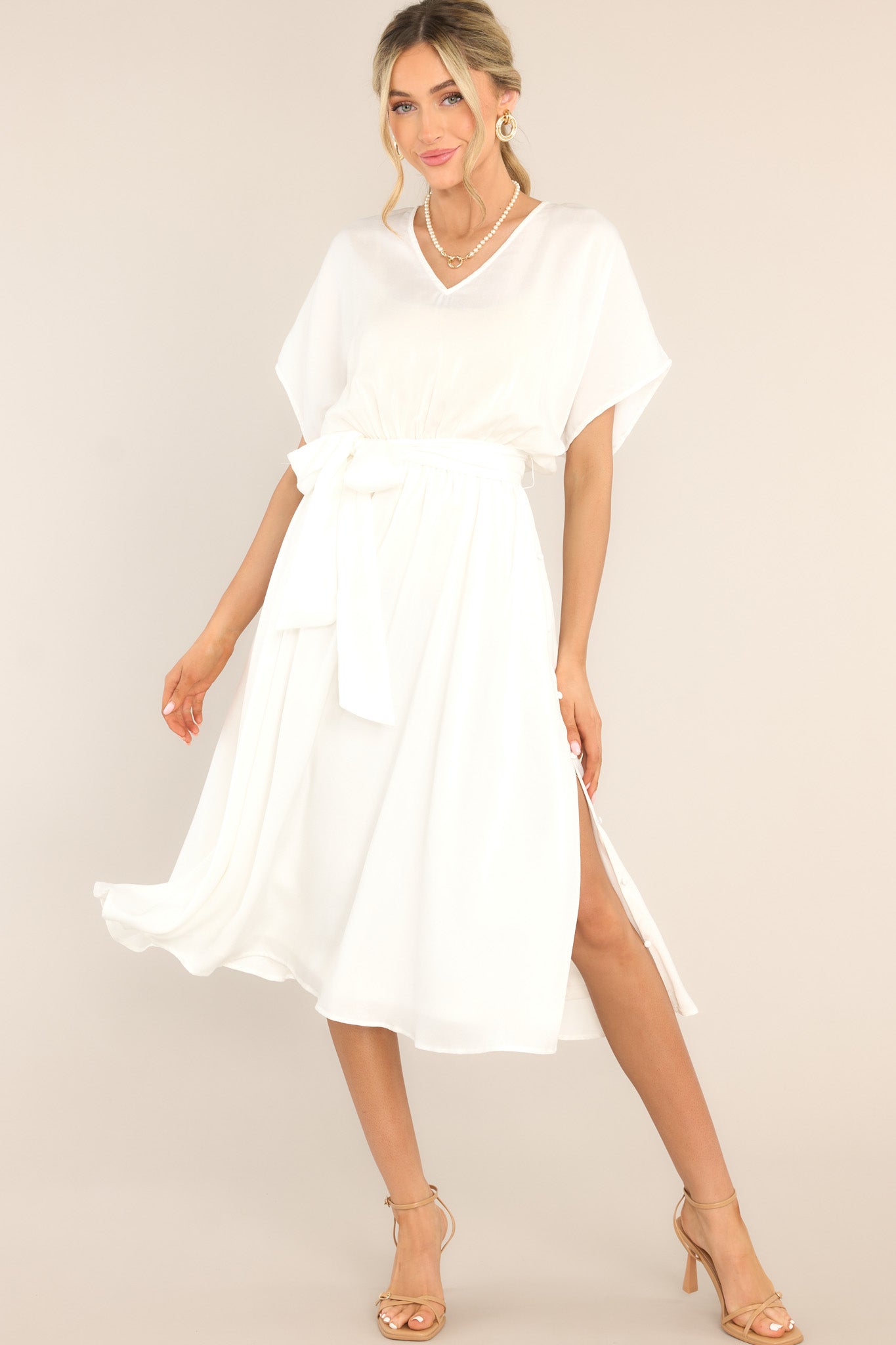 This all white midi dress features a V-neckline, an open back with a self-tie closure, flowy cap sleeves, an elastic waistband with a self-tie fabric belt, and button detailing down the side of the skirt.