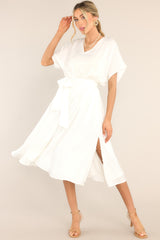 Full body view of white midi dress that features a V-neckline, cap sleeves, an elastic waistband with a self-tie fabric belt, and button detailing down the side of the skirt.