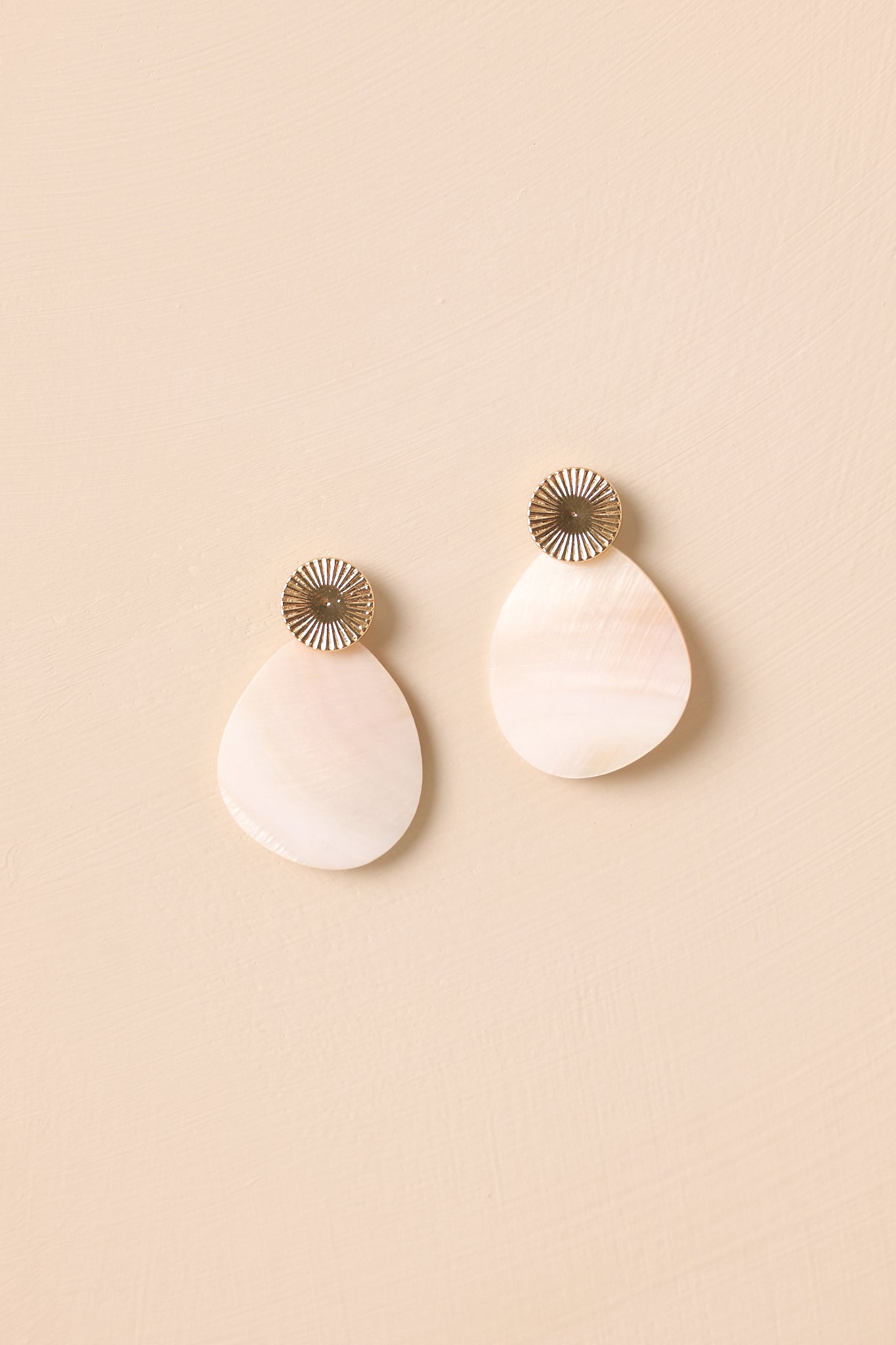 A shot of the gold textured stud earrings with ivory pearlescent drop charms displayed against a beige background.